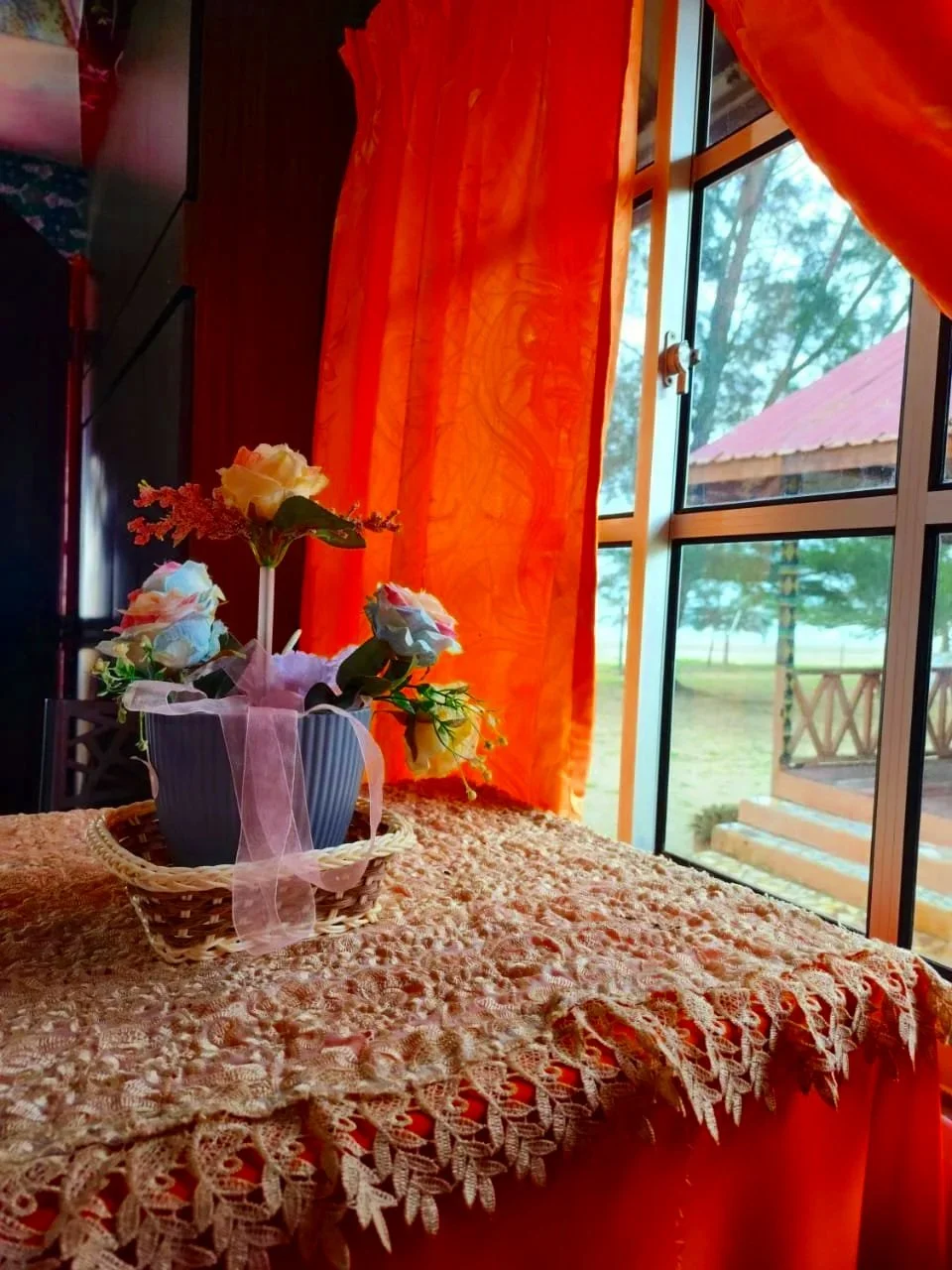 A table covered with a lacy tablecloth, holding a basket with a potted flower arrangement, positioned near a window with orange curtains, overlooking an outdoor patio and trees.