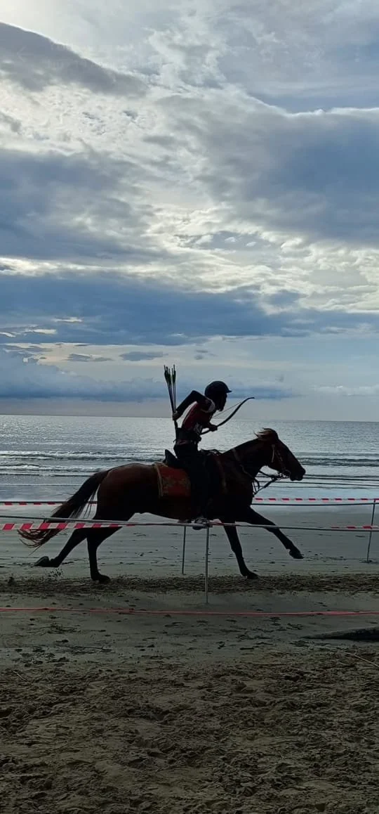 A person riding a horse along the beach at sunset, with the sky filled with clouds and the ocean in the background.