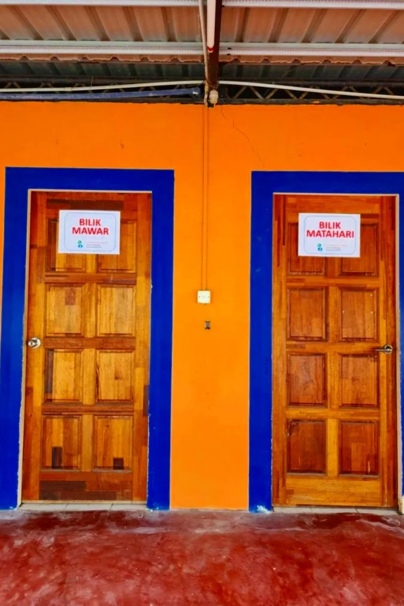 Two wooden doors with signs in Malay, one labeled 'Bilik Mawar' and the other 'Bilik Matahari,' surrounded by orange walls with blue trim, under a metal roof, and a cracked wall section.