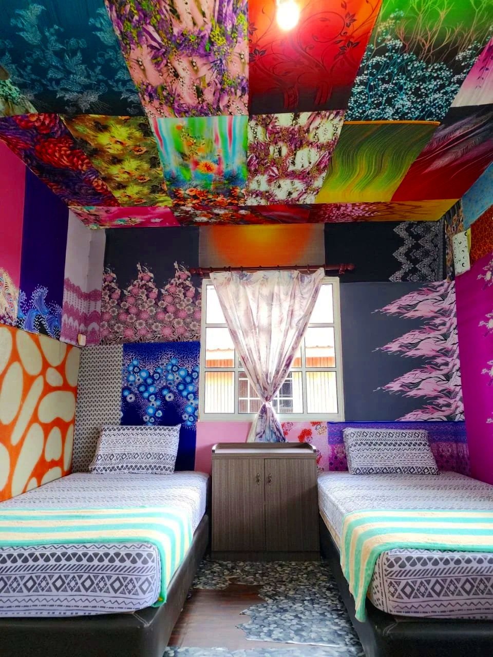 A bedroom with two single beds with striped bedding, a small wooden cabinet between them, colorful patterned walls, and a ceiling covered with a patchwork of vibrant floral and abstract fabric panels. There is a window with light-colored curtains and a patterned rug on the floor.
