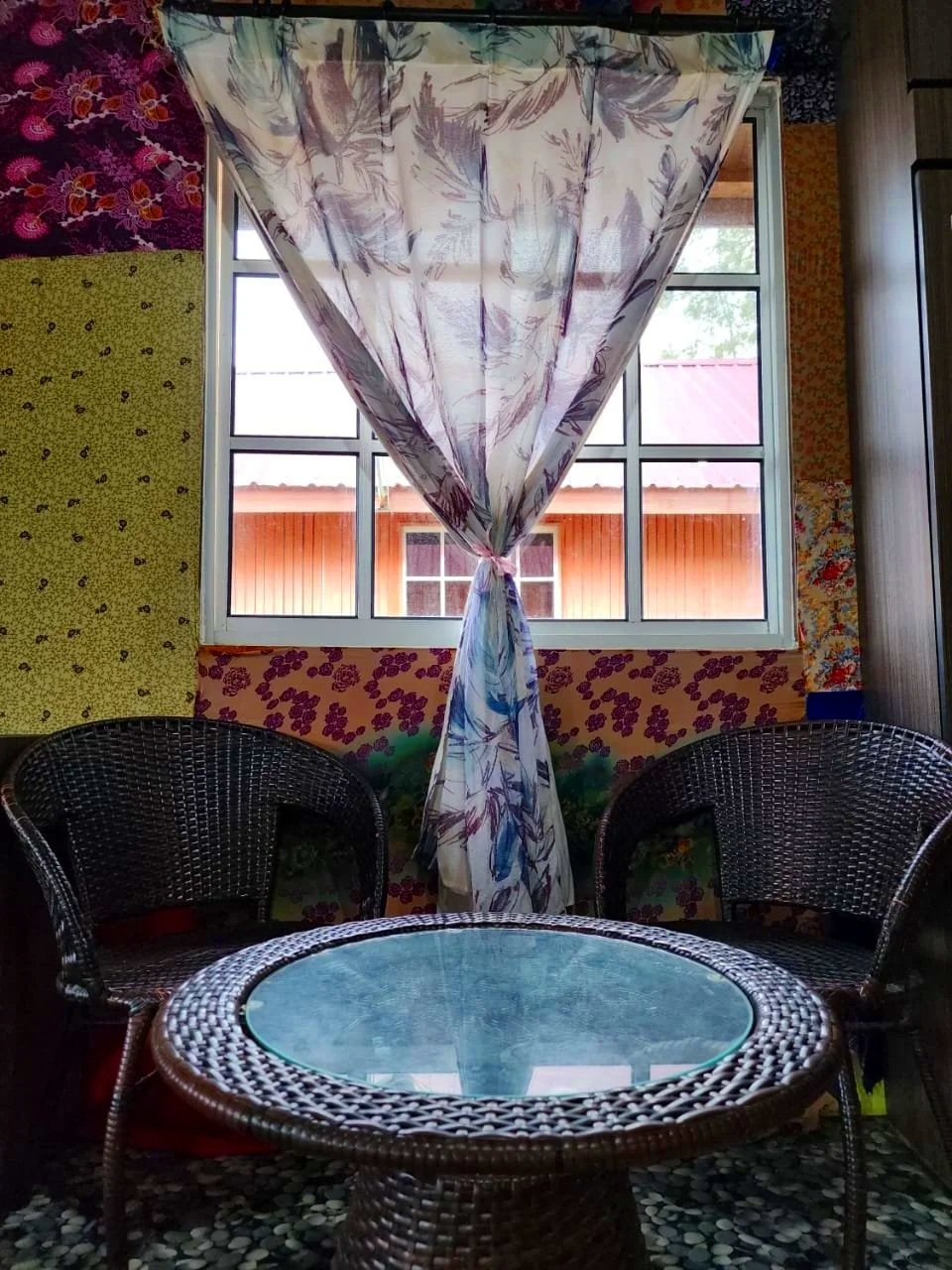 A cozy indoor seating area with a round wicker table with a glass top and two wicker chairs. A window with a floral curtain tied in the middle lets in natural light, with a view of a wooden building with a red roof outside.