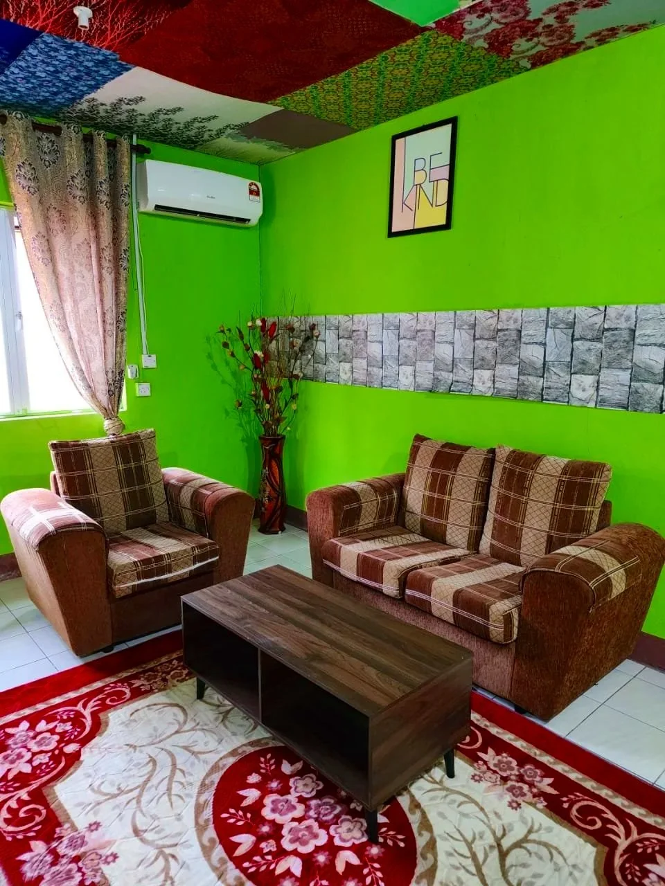 Living room with bright green walls, two plaid armchairs and a matching sofa, a wooden coffee table, floral area rug, window with pink patterned curtains, a vases with artificial branches, wall art, and a split Air Conditioner.
