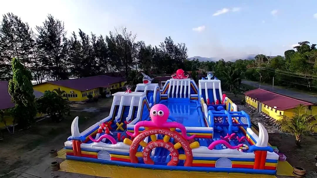 Colorful inflatable ocean-themed water park with slides, sea creature decorations, and a welcome sign, set outdoors near trees and buildings.