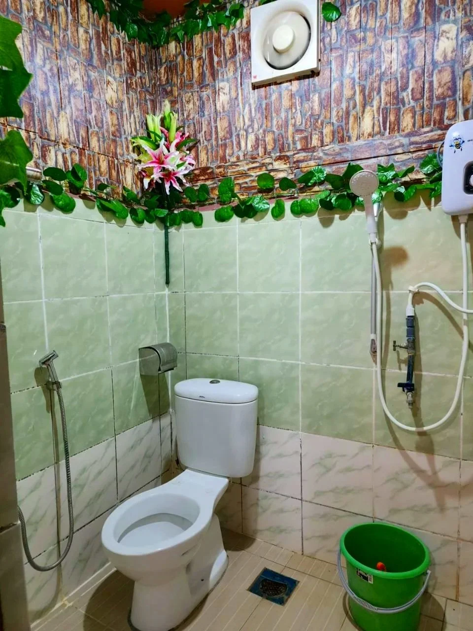 A bathroom with a toilet, a green bucket, and wall-mounted toiletry fixtures. Decor includes artificial greenery and pink lilies on the upper part of the wall. The walls are tiled with green and beige tiles, and there is an air ventilation unit on the upper wall.