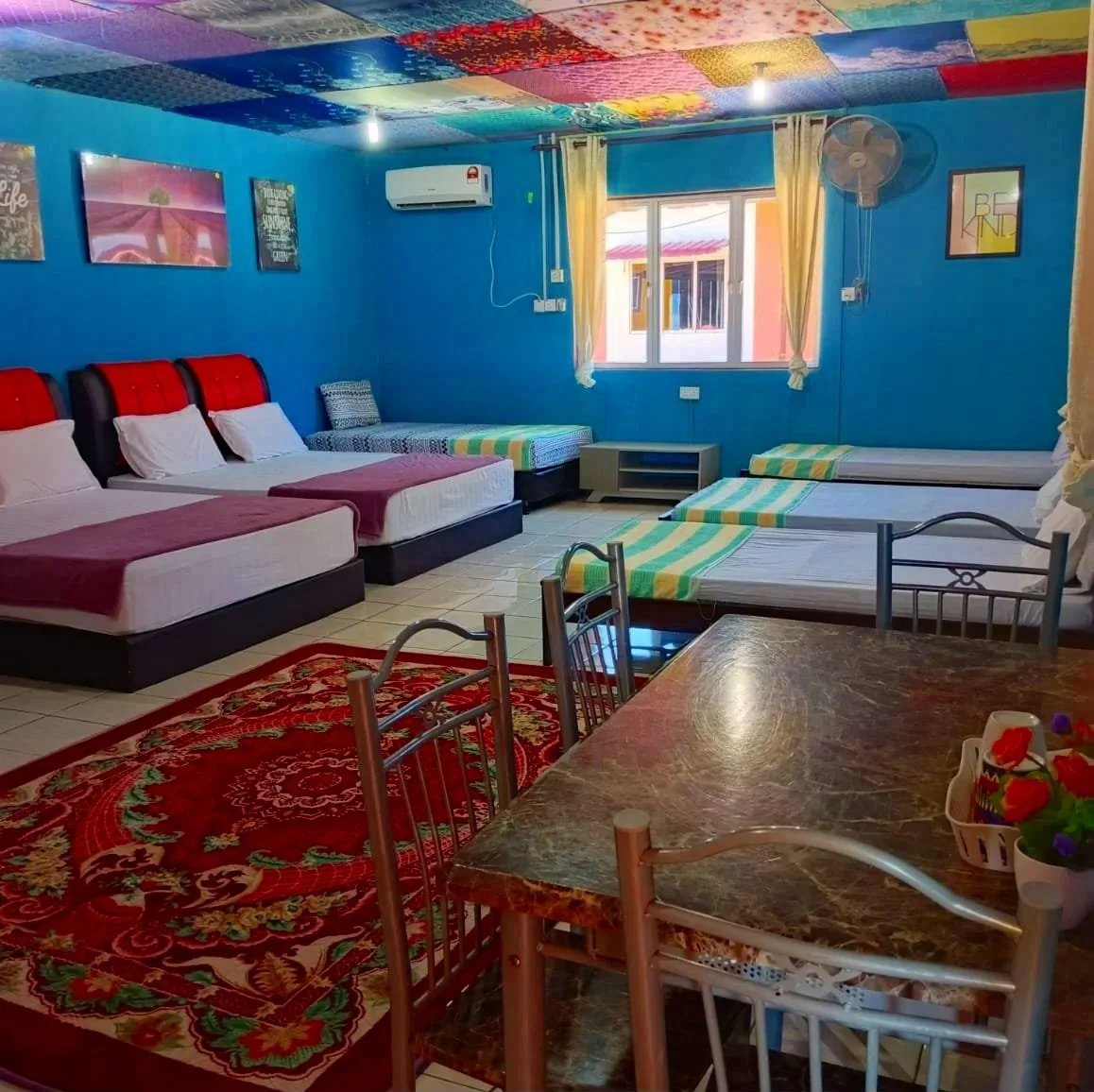 Colorful bedroom with blue walls and a textured ceiling, featuring multiple beds with striped and solid bedding, a window with yellow curtains, a red patterned rug, a marble-top table with metal chairs, and wall art.