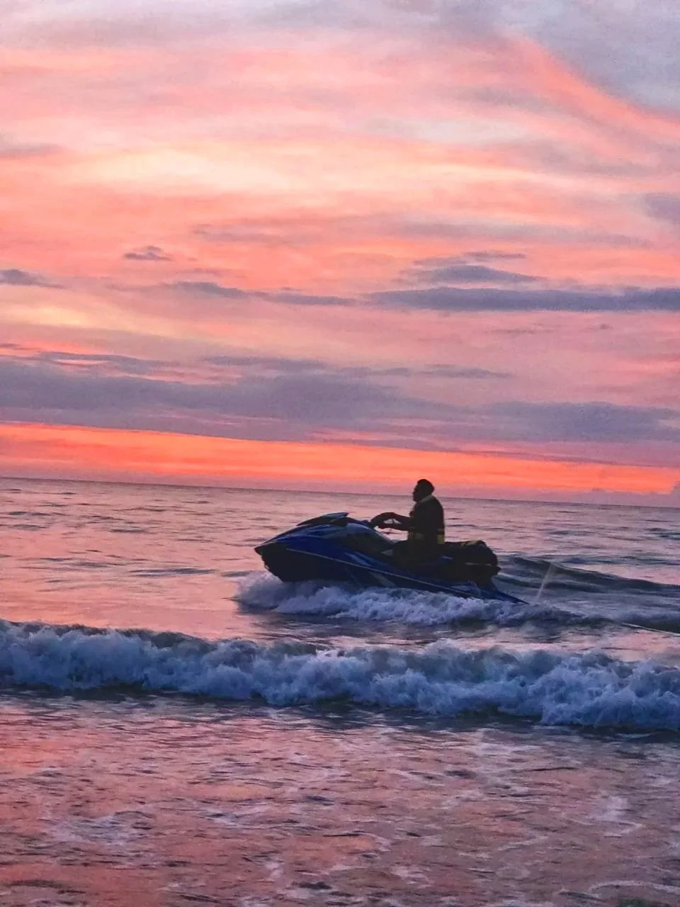 Person riding a jet ski on the ocean during a colorful sunset with pink, purple, and orange clouds