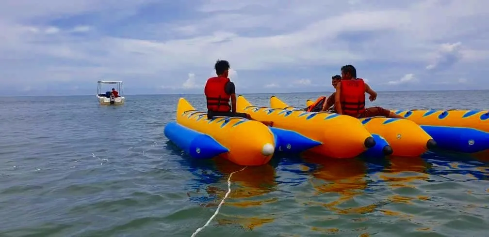 Three people on an inflatable banana boat in the ocean with a boat in the distance under a partly cloudy sky.