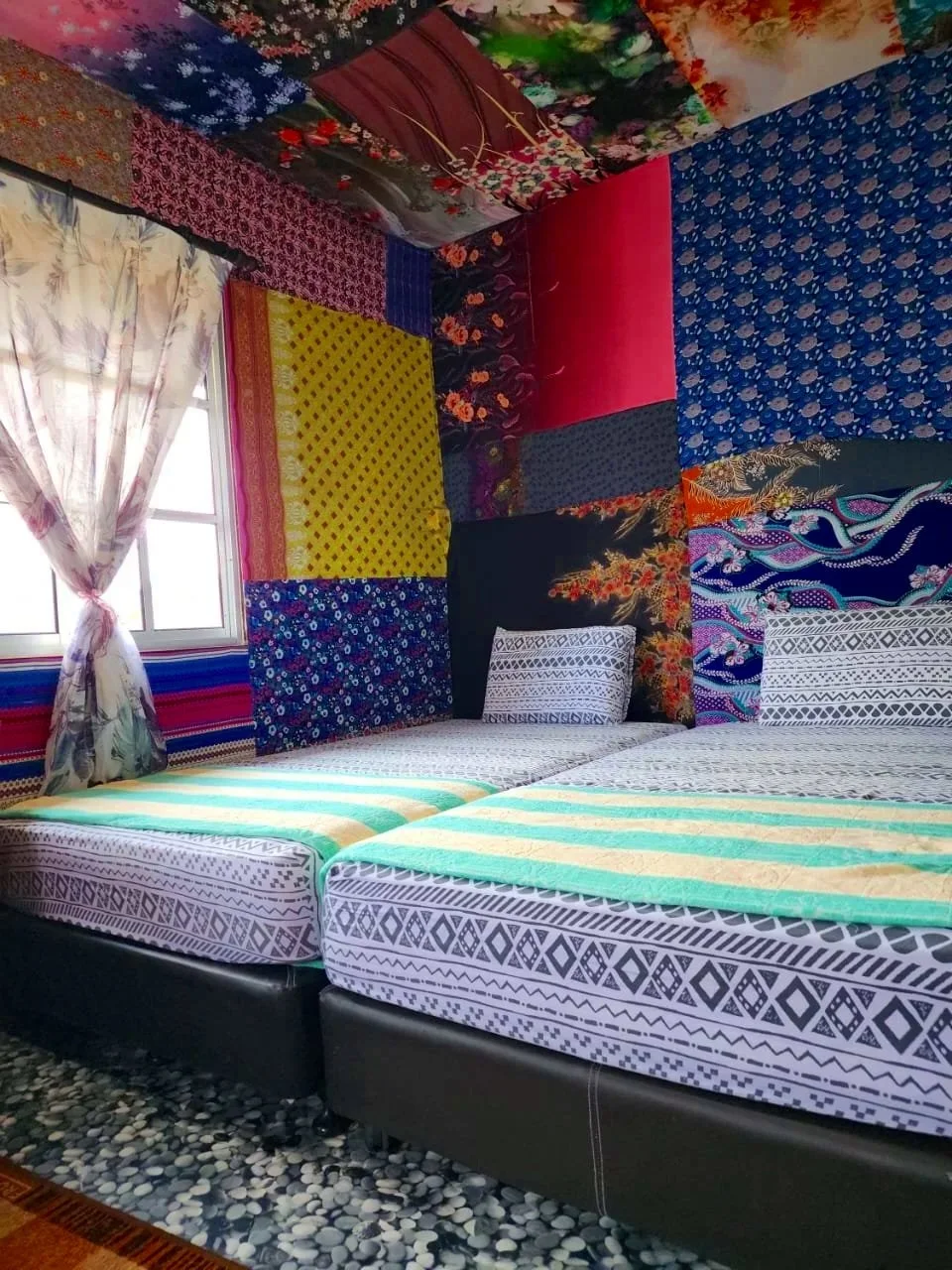 A bed with patterned sheets and pillows in a room decorated with colorful, patchwork fabric wall coverings and ceiling, with a window having floral curtains.