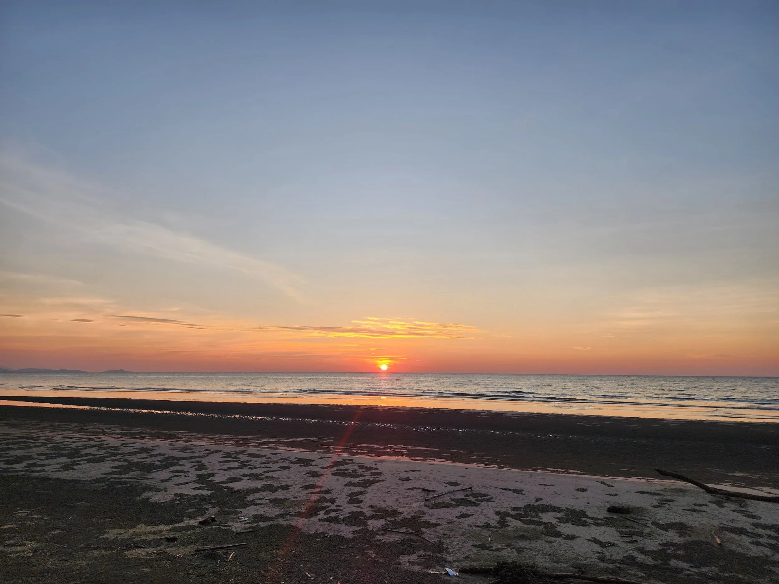 Sunset over the ocean with a clear sky and some clouds, viewed from a sandy beach.