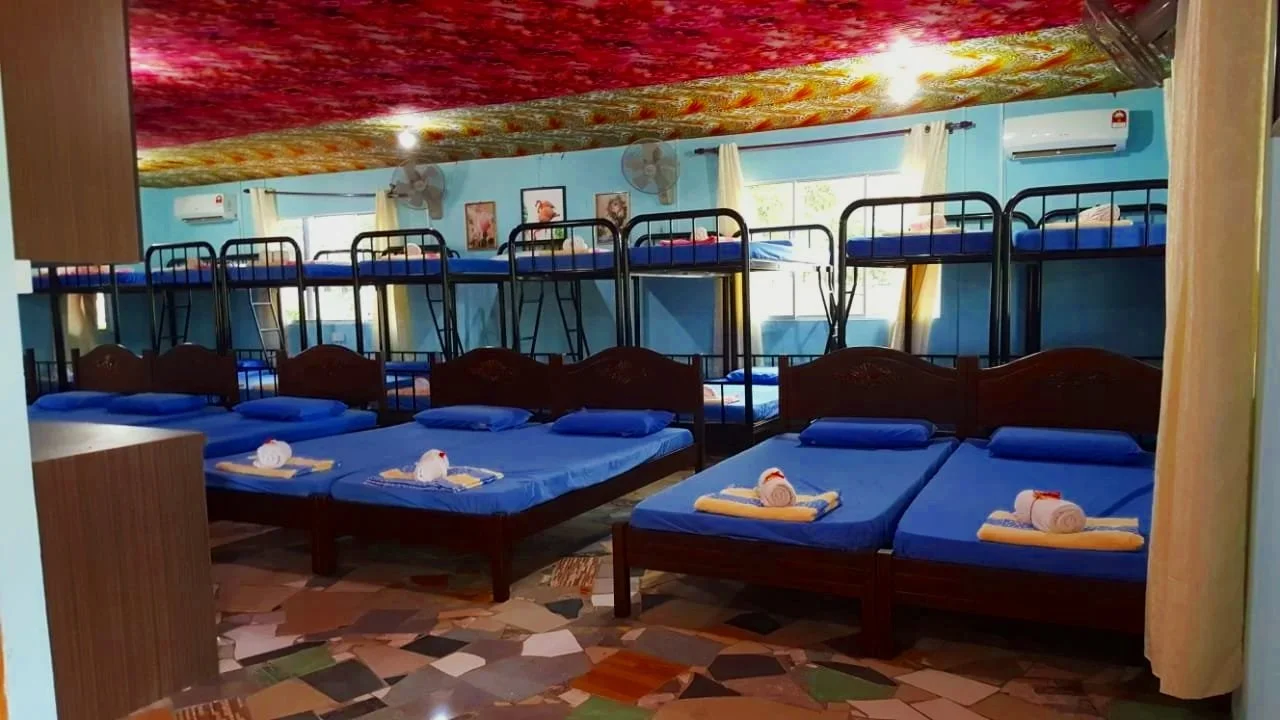 A room with multiple beds, including three single beds in the foreground each with blue bedding and towels, and several bunk beds in the background, all with blue bedding. The room has light blue walls, framed pictures, windows with white curtains, and a patterned floor.