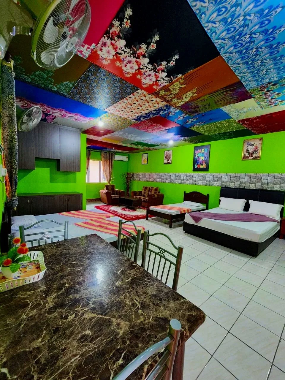 A colorful room with a decorated patchwork ceiling and walls painted bright green. The room contains a bed, a small mattress, a sitting area with two sofas, and a marble dining table with chairs. There are wall pictures, a window with curtains, and a ceiling fan.