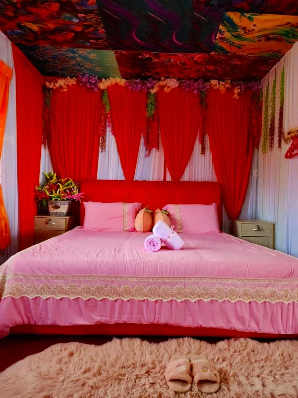 A bedroom decorated with pink and red elements, featuring a pink bed with lace trim, pink pillows, and rolled towels with flowers on the bed. Red drapes and floral arrangements adorn the room, with a floral ceiling and a plush pink rug in front of the bed.