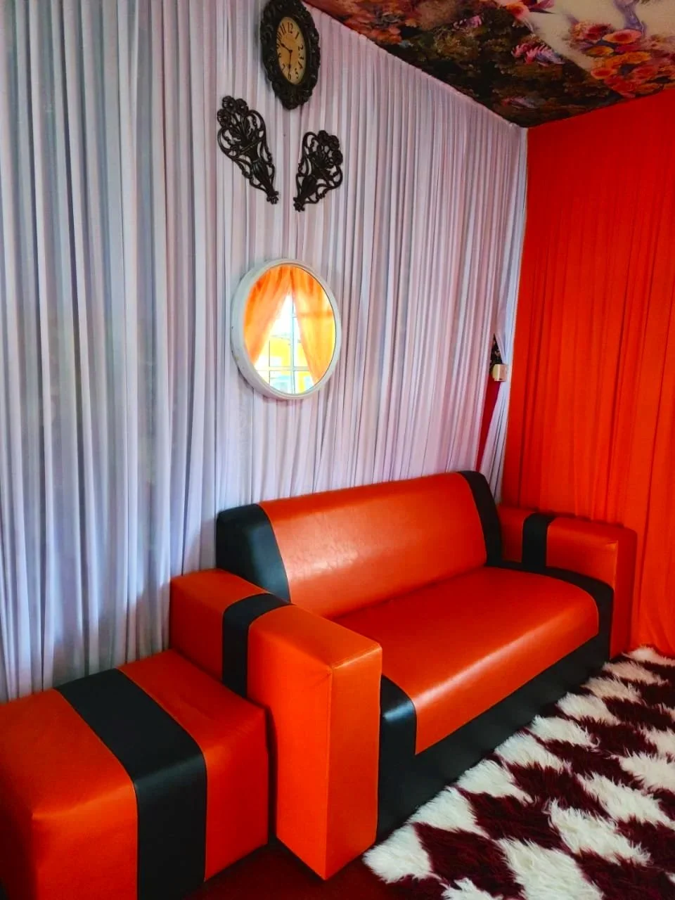 A vibrant living room with bold, colorful decor. There is a red and black leather sofa, a black and white patterned rug, and a wall of striped white and grey curtains. The walls are painted red, and the ceiling has a floral pattern. A round mirror hangs on the wall, and a vintage clock along with ornamental black wall art are displayed above the mirror.