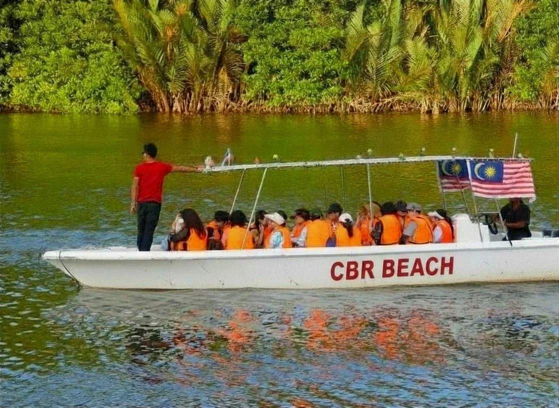 A boat with people wearing orange life vests on a river, labeled 'CBR BEACH' with Malaysian flags, and a person standing at the front guiding the boat.