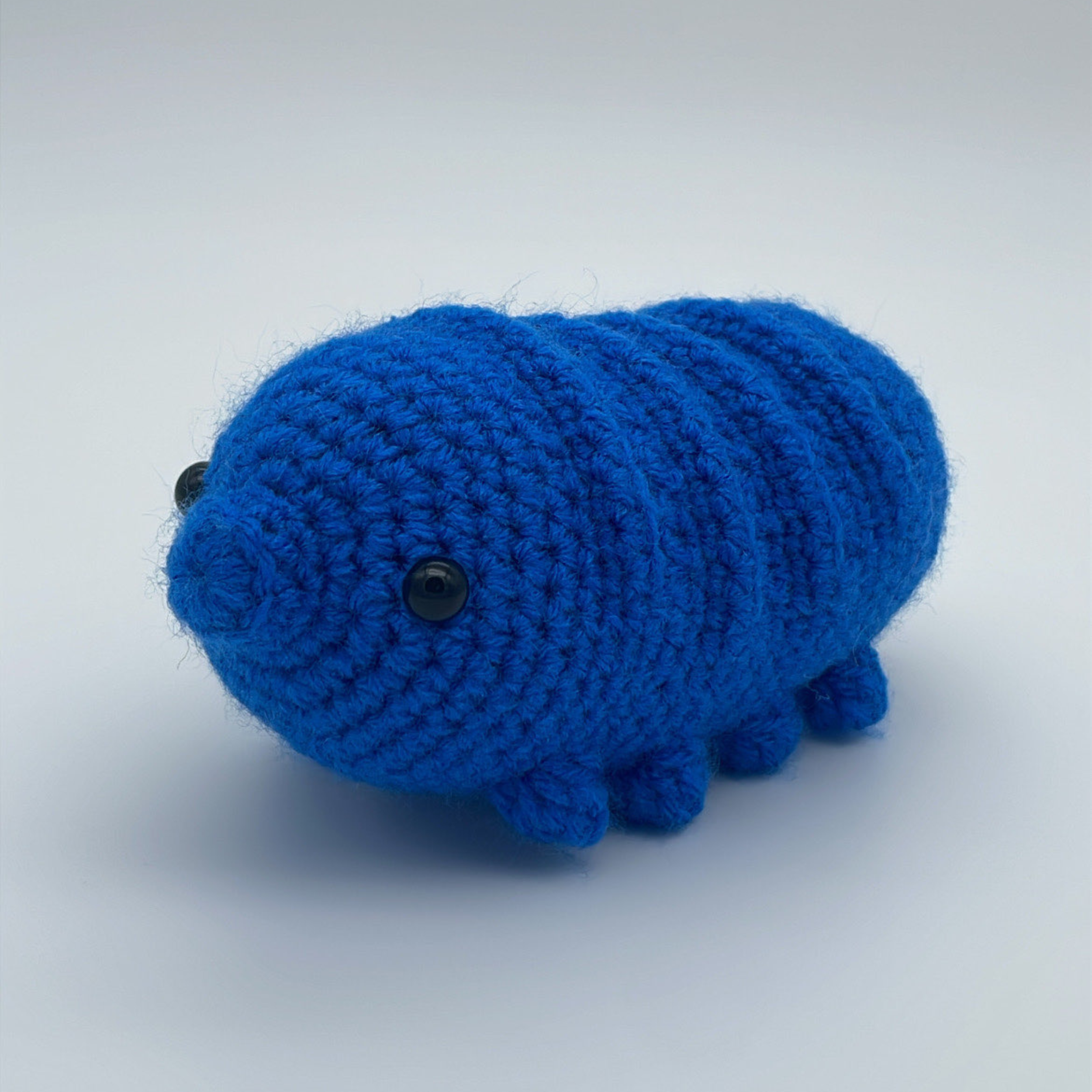 Blue crocheted tardigrade plush with a rounded segmented body, small black bead eyes, and short stubby legs, photographed against a light background.