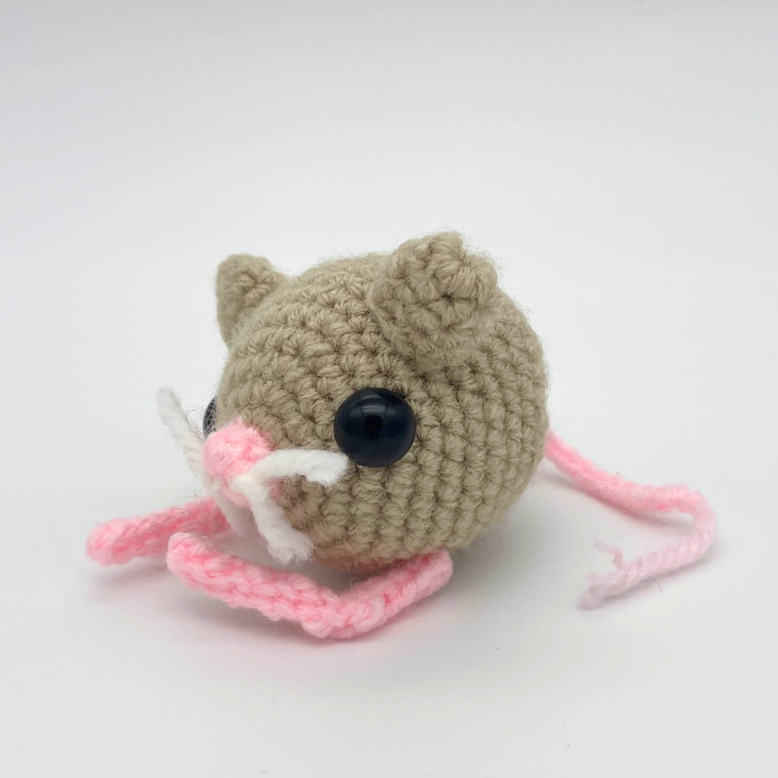 Small crocheted pygmy jerboa plush with a tan round body, large black bead eyes, small ears, pink nose, white whiskers, and long pink tail, photographed against a light background.