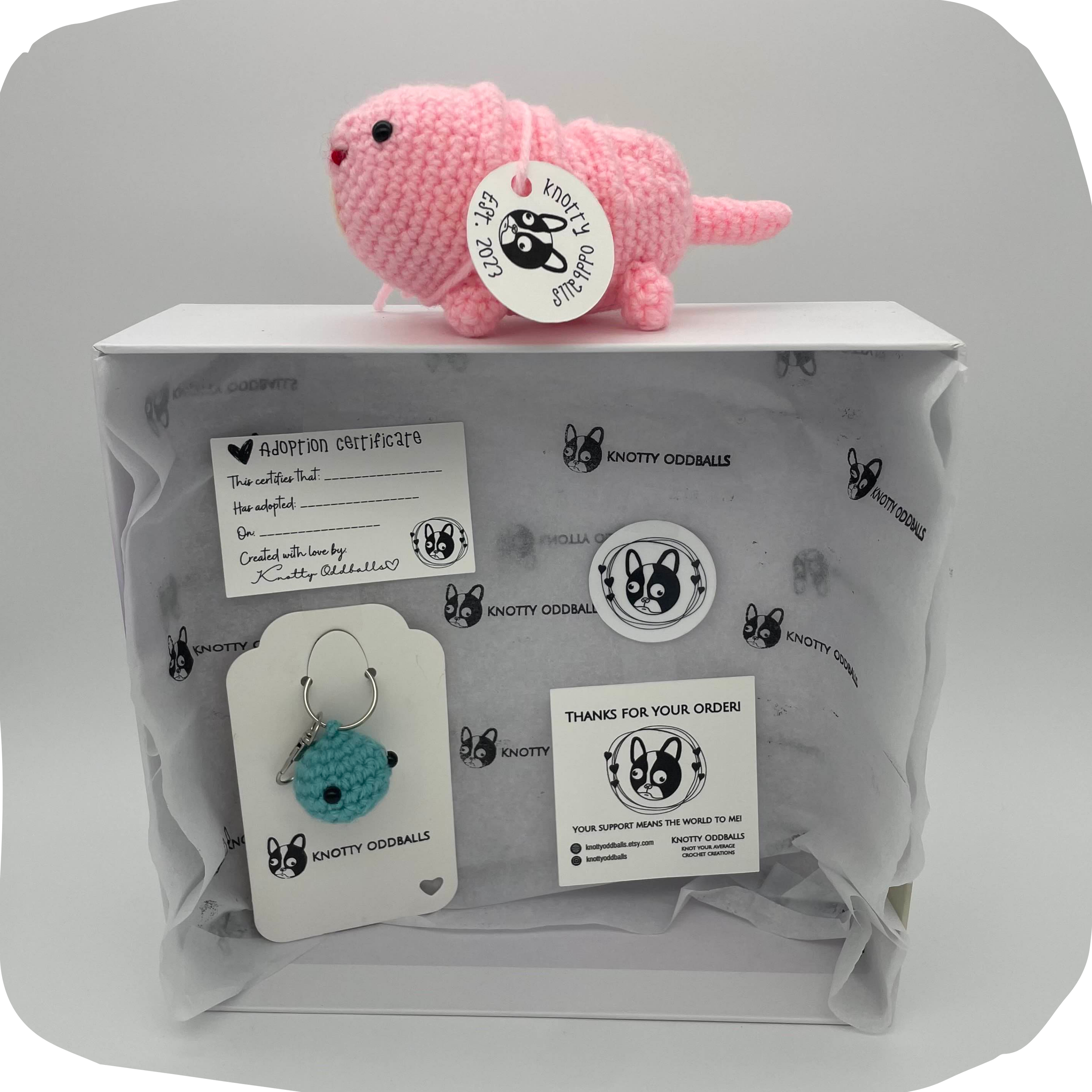 Crochet adoption kit featuring a pink mole rat plush with a small collar, displayed on top of a wrapped package with branded paper, alongside a blue crocheted keychain, printed adoption certificate, sticker, and thank you card.