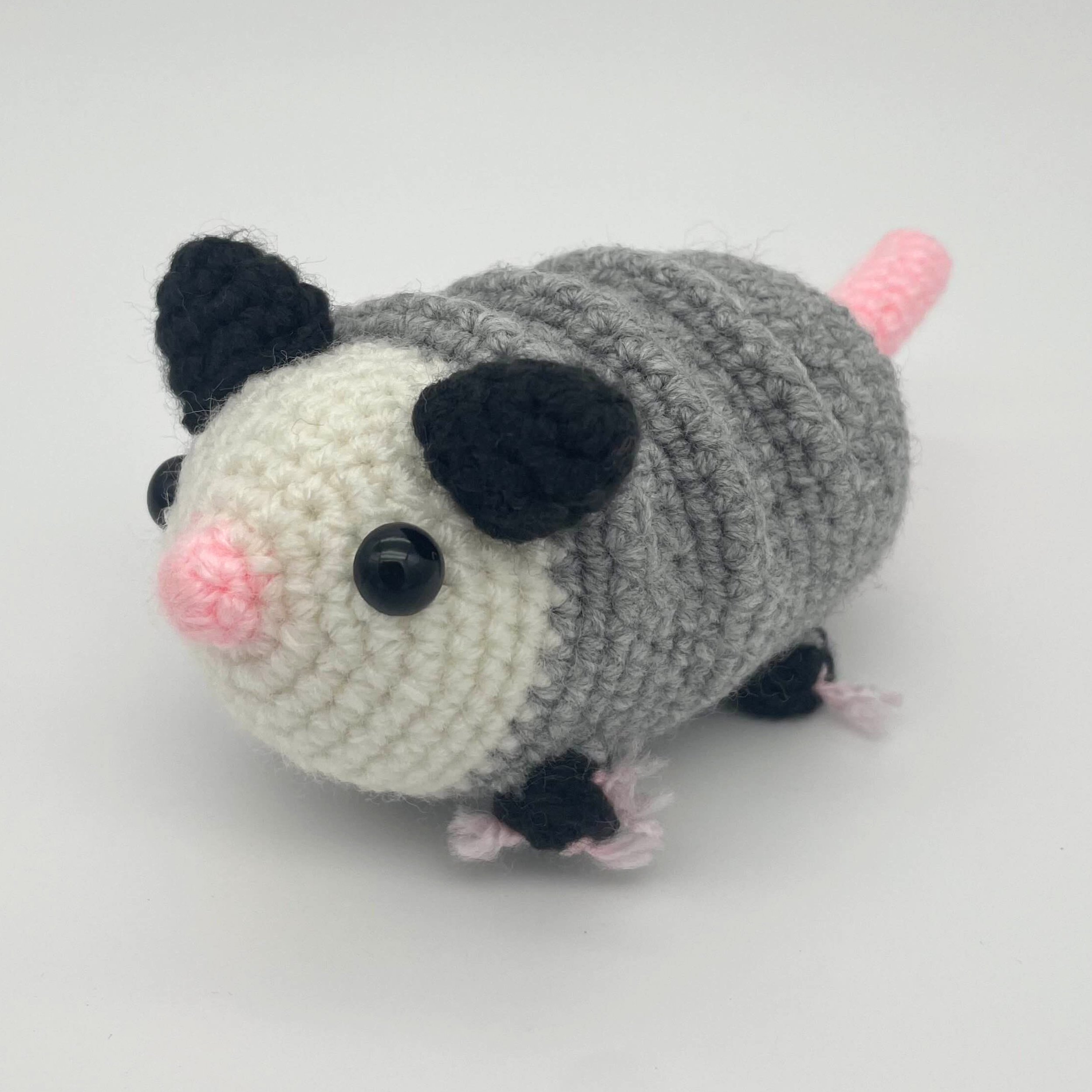 Gray and white crocheted opossum plush with a rounded body, black ears, black bead eyes, pink nose, small legs, and a pink tail, photographed against a light background.