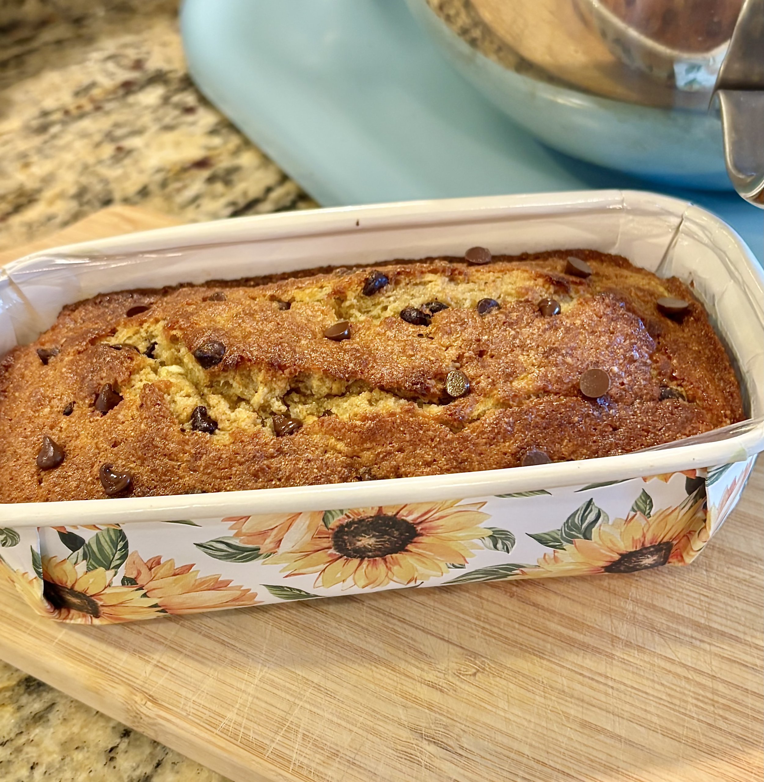 Close-up of a freshly baked bread or cake with chocolate chips on top, placed in a decorative container with sunflower and leaves design, on a wooden surface.