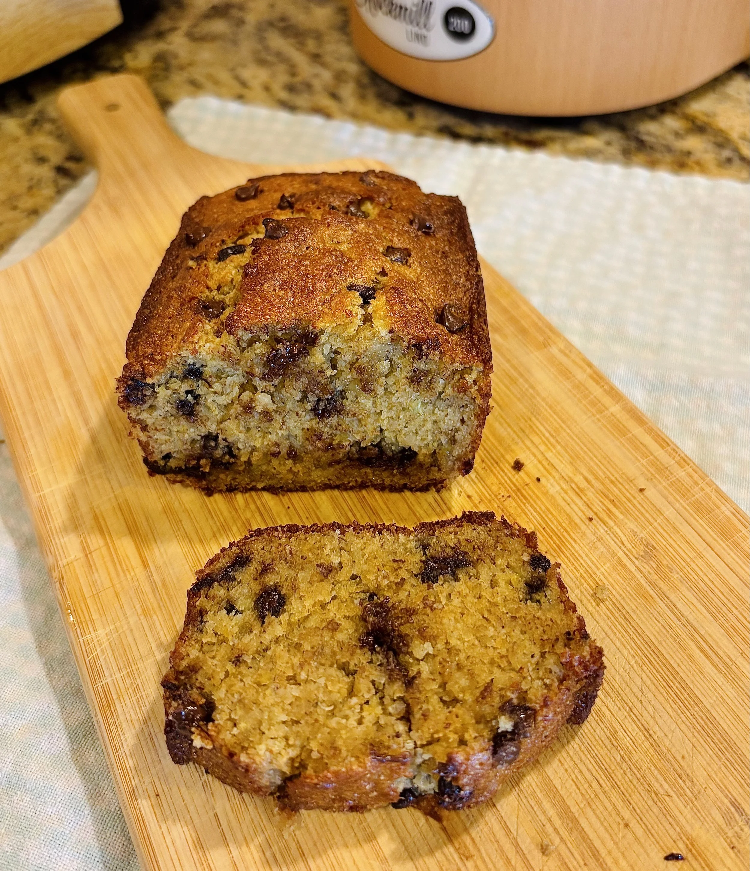 Close-up of a slice of homemade banana bread with chocolate chips on a wooden cutting board, with the rest of the loaf in the background on a granite kitchen countertop.