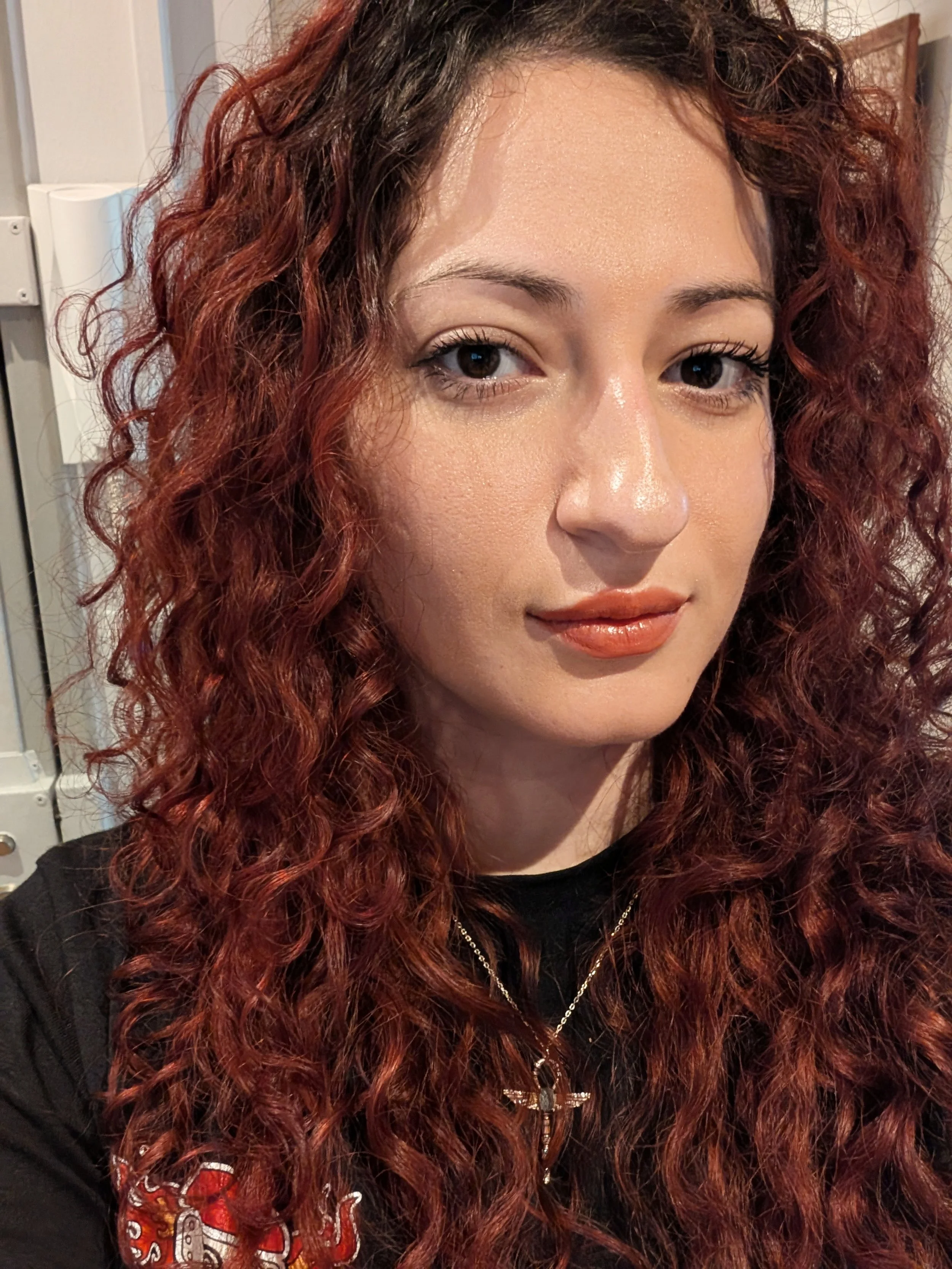 Un portrait d'une femme aux cheveux longs, bouclés et rouge foncé, portant un sweat noir avec un motif rouge et blanc sur le bras gauche, et des bijoux dorés, dans un intérieur.