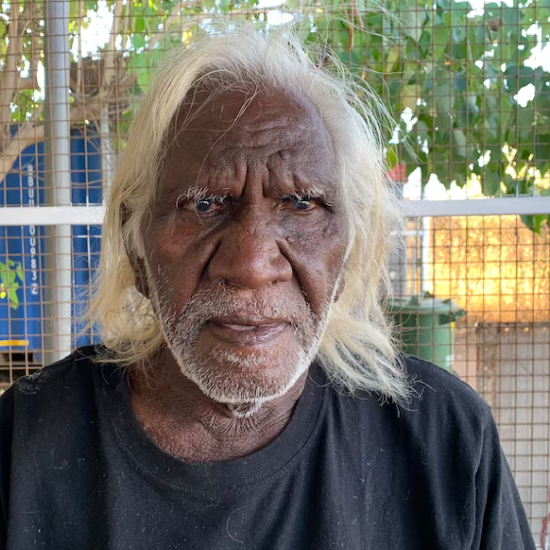 Close-up of an elderly Indigenous man with long white hair and beard, wearing a black shirt, outdoors in front of a wire fence with green foliage behind him.