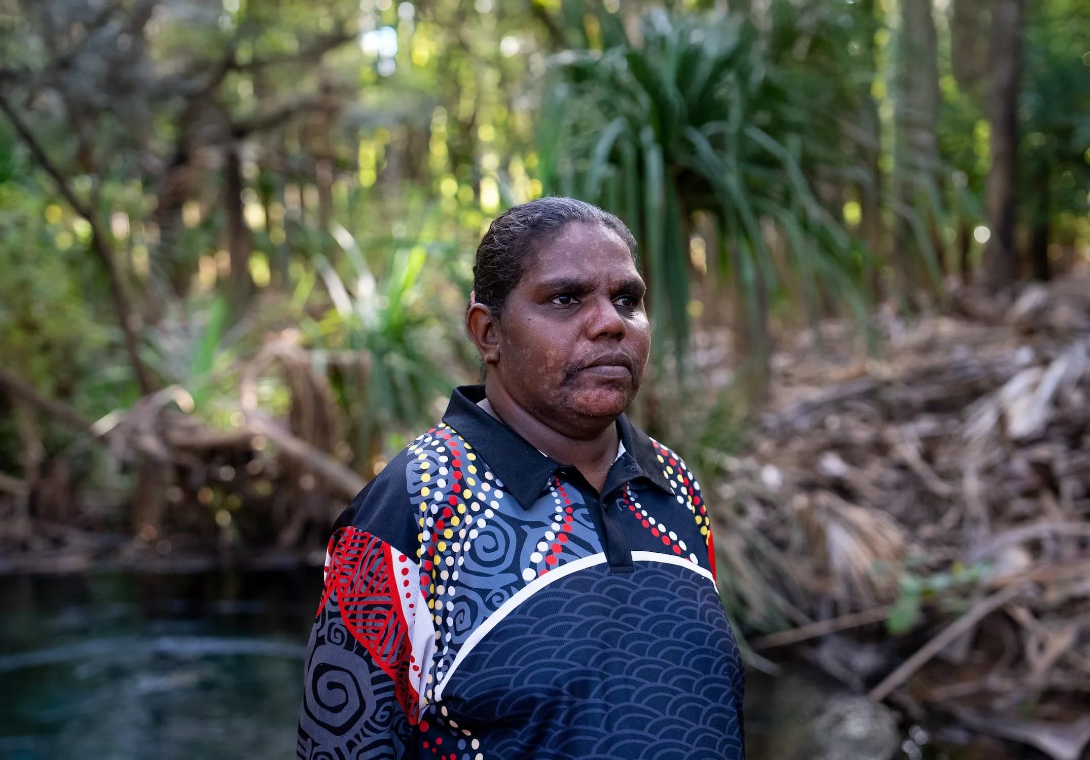 An indigenous woman with dark hair and brown skin standing outdoors in a lush, green forest.