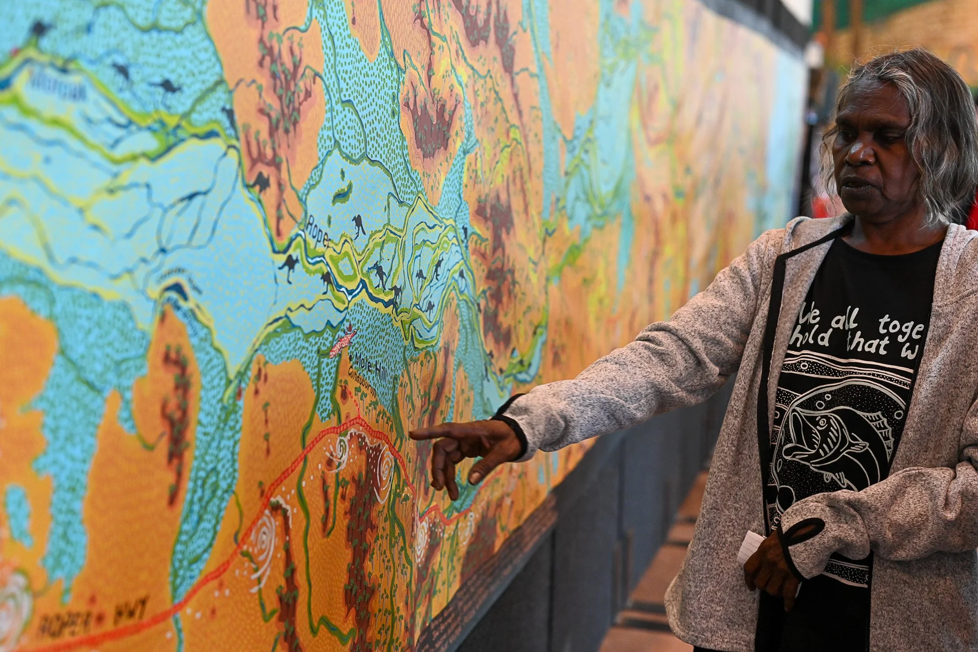 An elderly person with gray hair is pointing at a large, colorful map on a wall, discussing or examining it. The map includes waterways, mountains, and various geographical features, with labels and detailed illustrations.