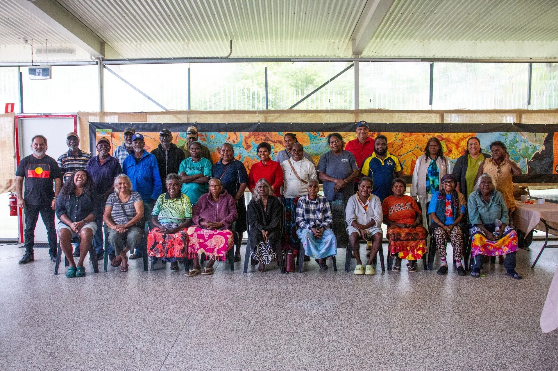 Group of diverse people of various ages posing for a photo indoors, with a colorful map or mural behind them. Some are seated and some are standing, smiling at the camera.