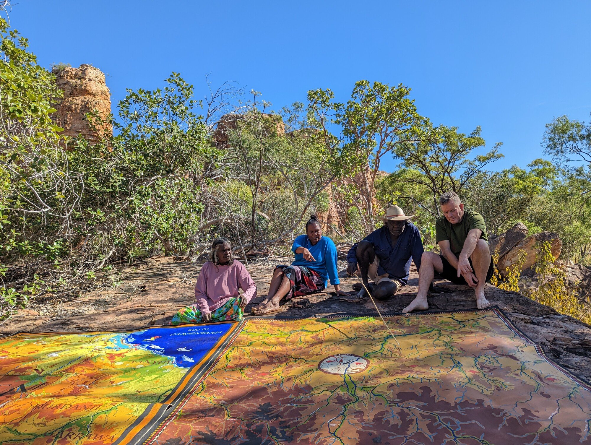 Indigenous and non-Indigenous people sit on a rock, looking over a large painted map of the Roper River