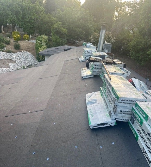 Roof shingles installation in progress with stacks of shingles and a hammer on a house roof, surrounded by trees and greenery.
