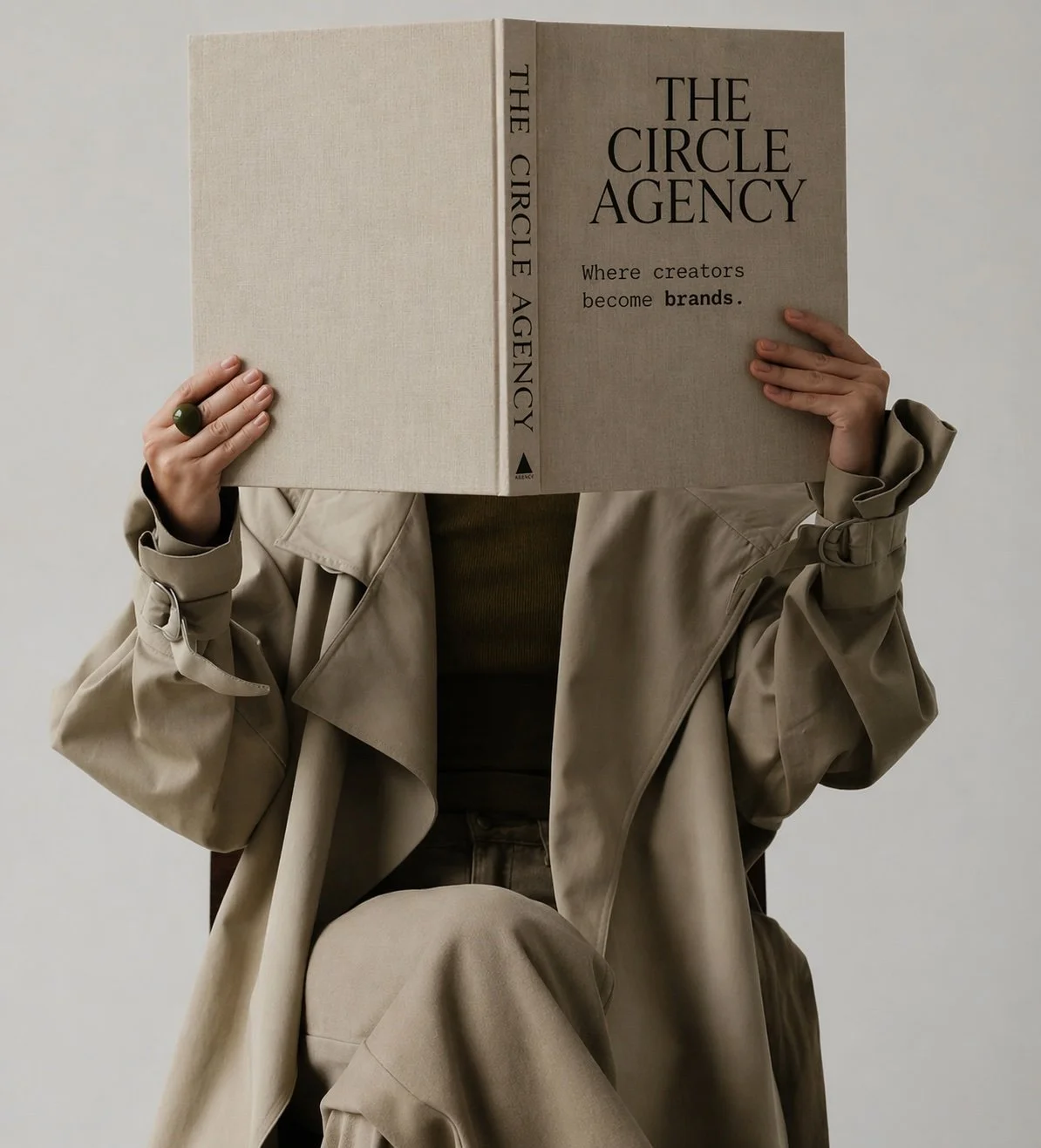 Person wearing a beige trench coat sitting and holding a large book titled 'The Circle Agency' in front of their face, with the cover reading 'Where creators become brands.'