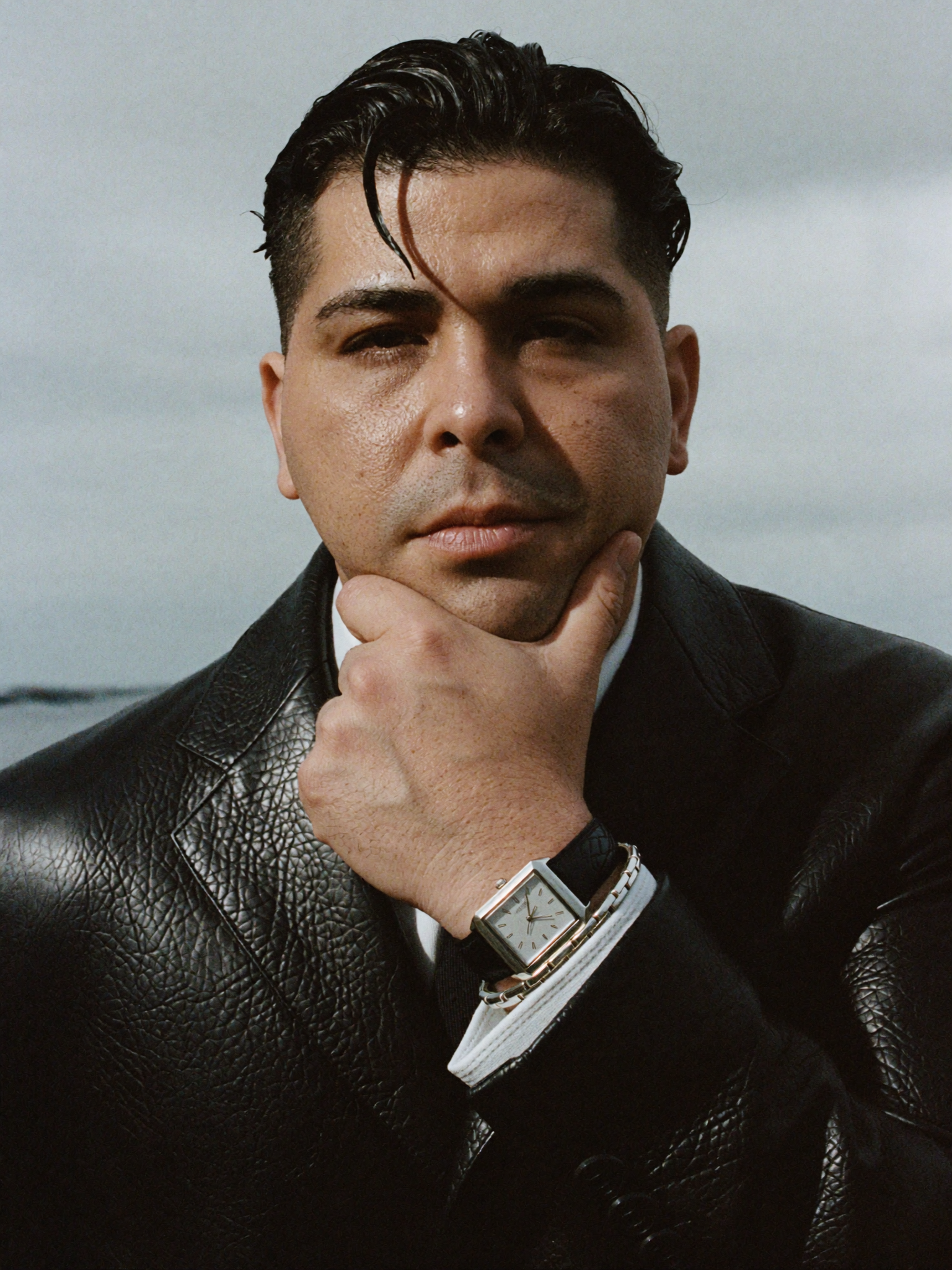 A man with dark, slicked-back hair and light skin, wearing a black leather jacket and a silver wristwatch, poses outdoors, resting his chin on his hand.