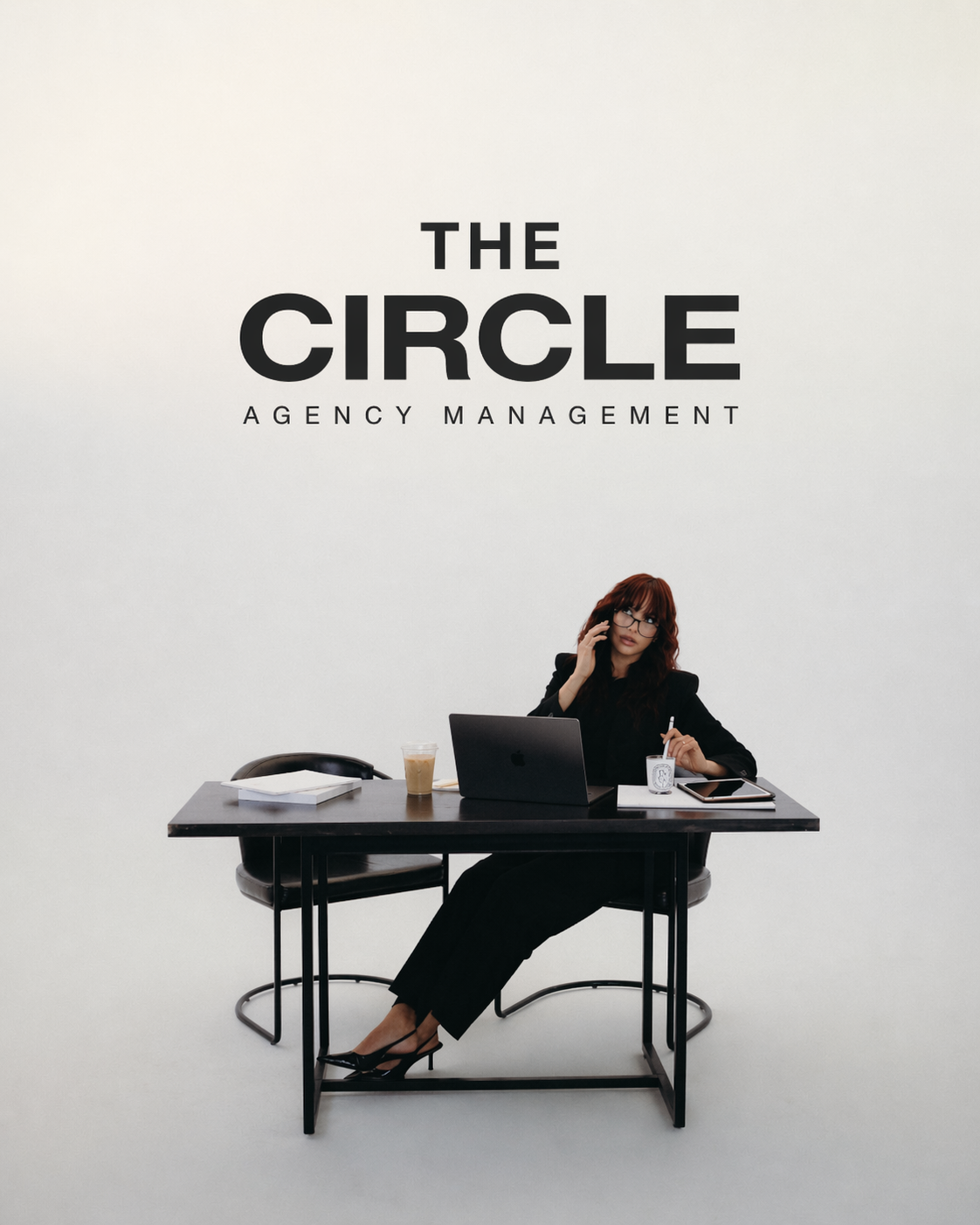 A woman with red hair and glasses sitting at a black desk, talking on the phone, with a laptop, coffee, and other office items, in front of a plain light background with the text 'The Circle Agency Management' above her.