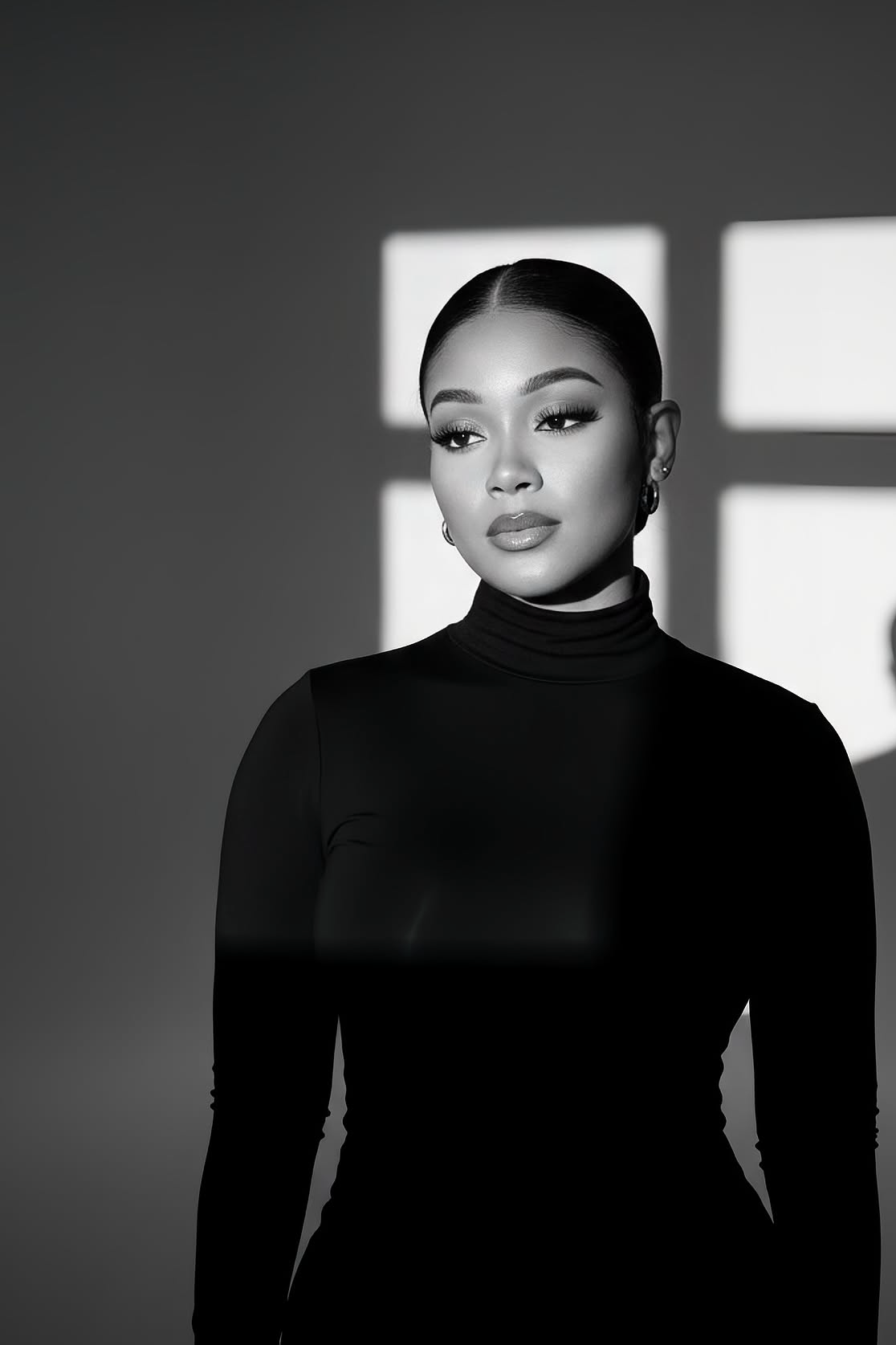 Black and white photo of a confident woman with dark hair slicked back, wearing a high-neck black top, standing indoors with sunlight casting a cross-shaped shadow on the wall behind her.