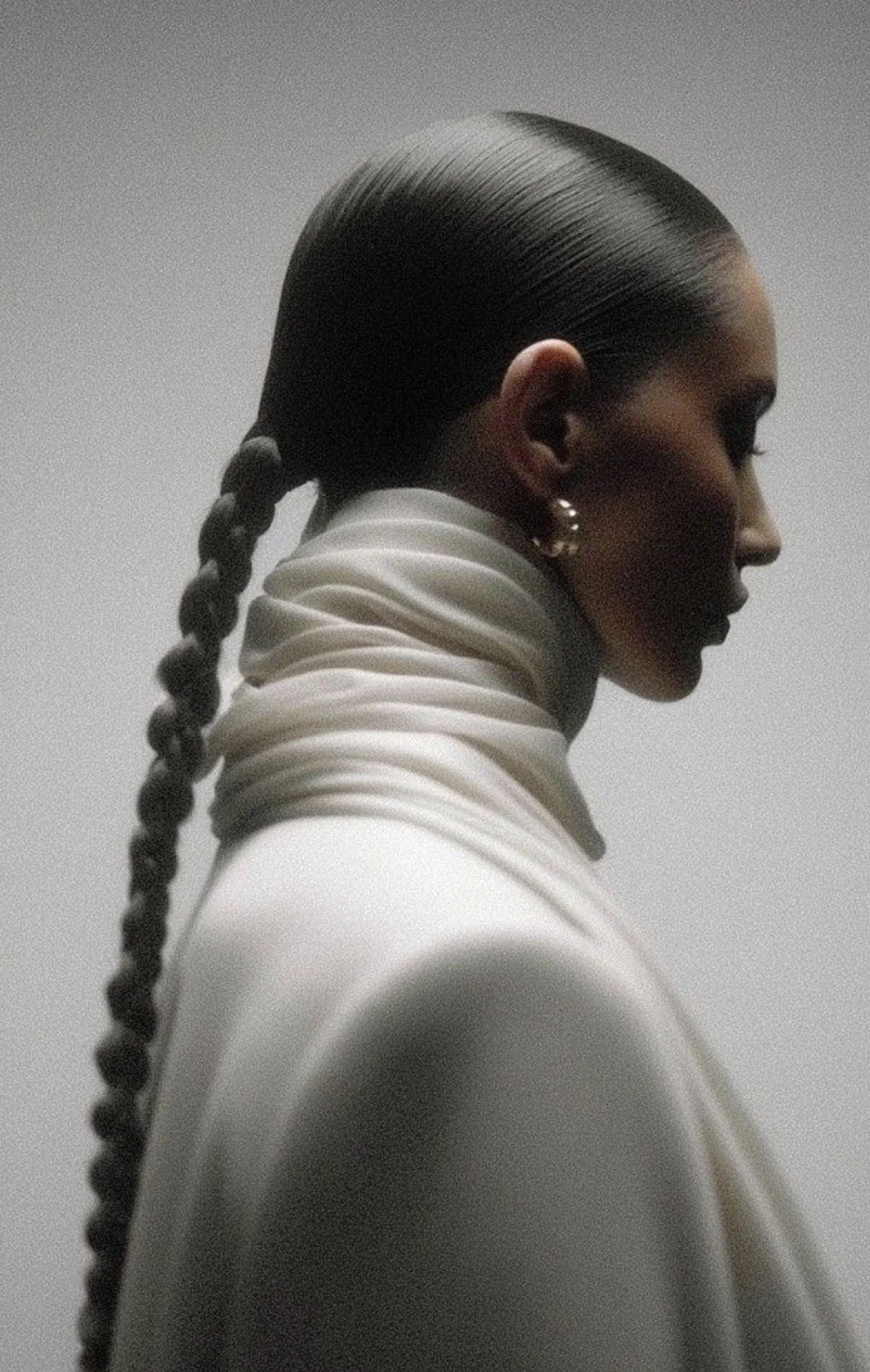 Side profile of a woman with slicked back hair in a braid, wearing a white high-neck top with ruffled details and pearl earrings.