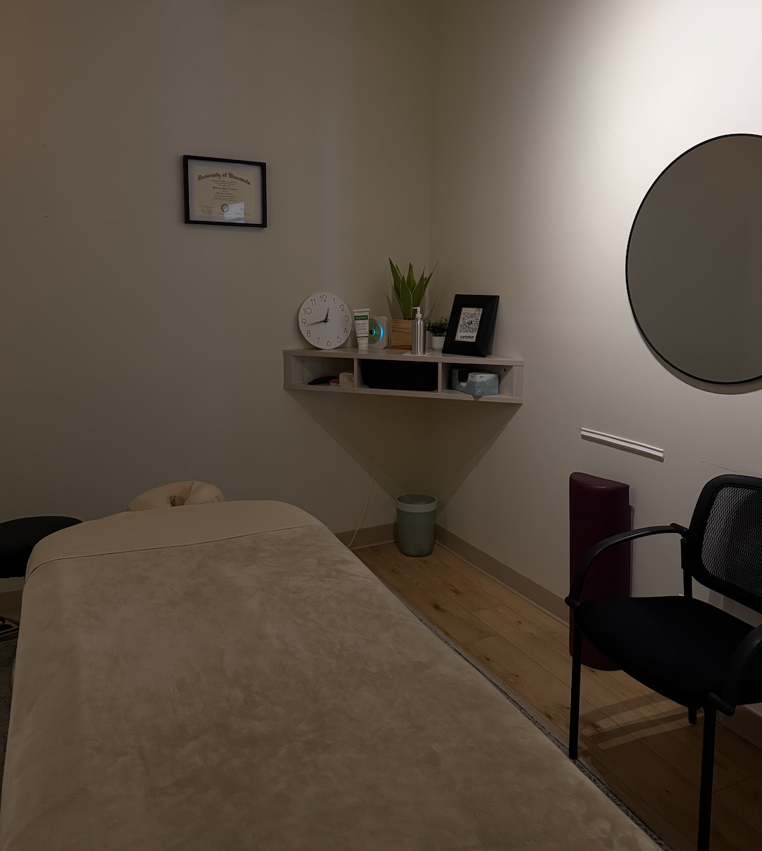 A massage therapy room with a bed covered in a beige sheet, a black chair, a floating shelf with a clock, plants, picture frame, and toiletries, and a framed diploma on the wall.