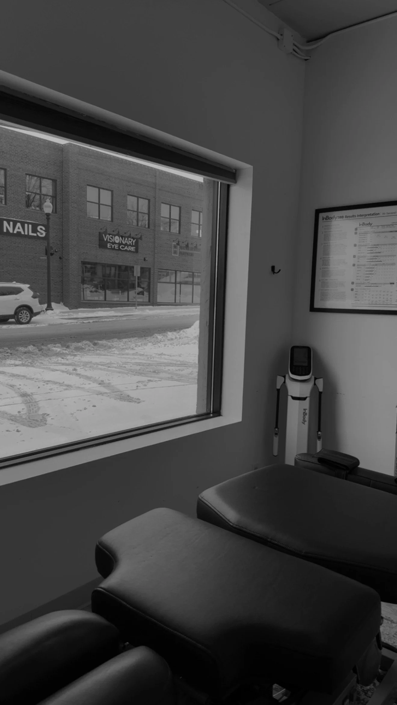 View from a window inside a medical or therapy office showing a waiting area with black leather chairs, a wall-mounted diagnostic device, and a framed chart. Outside, a snowy street scene with parked cars and commercial buildings.