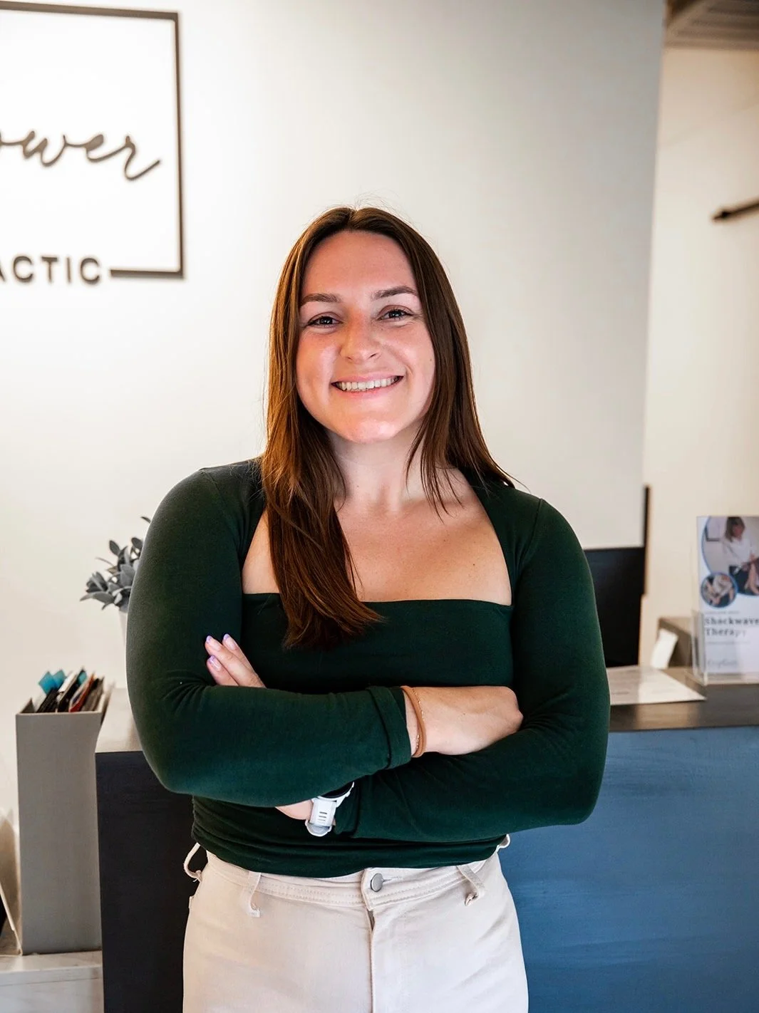 A woman with long brown hair and fair skin standing with arms crossed, smiling in a reception area or office. She is wearing a dark green off-shoulder top and light-colored pants.
