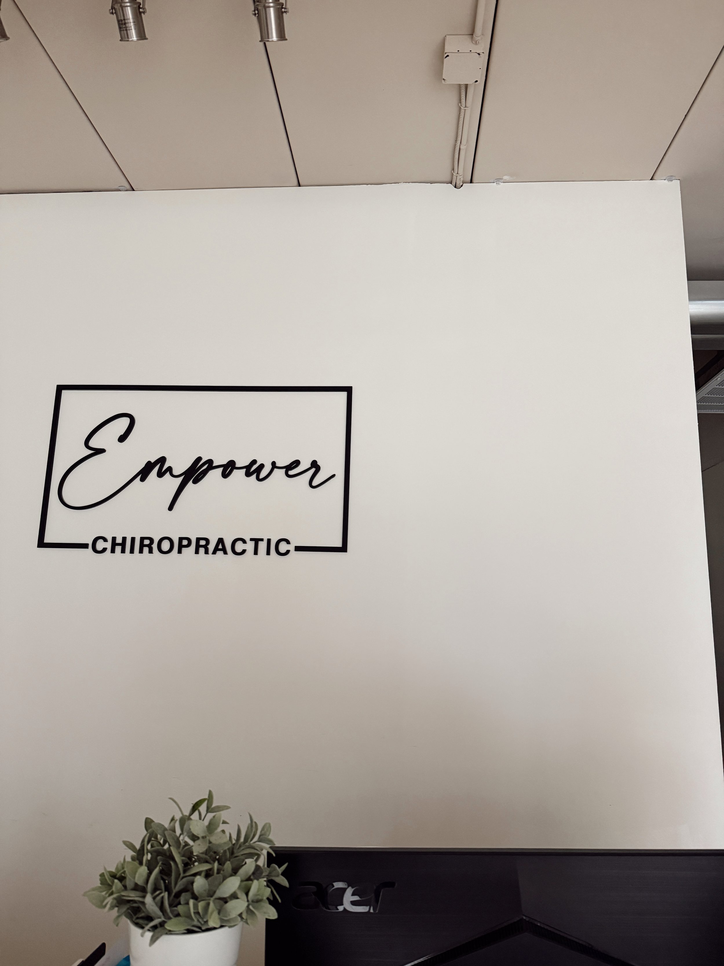 Office wall with a sign that reads 'Empower Chiropractic', a potted plant, and part of a computer monitor.