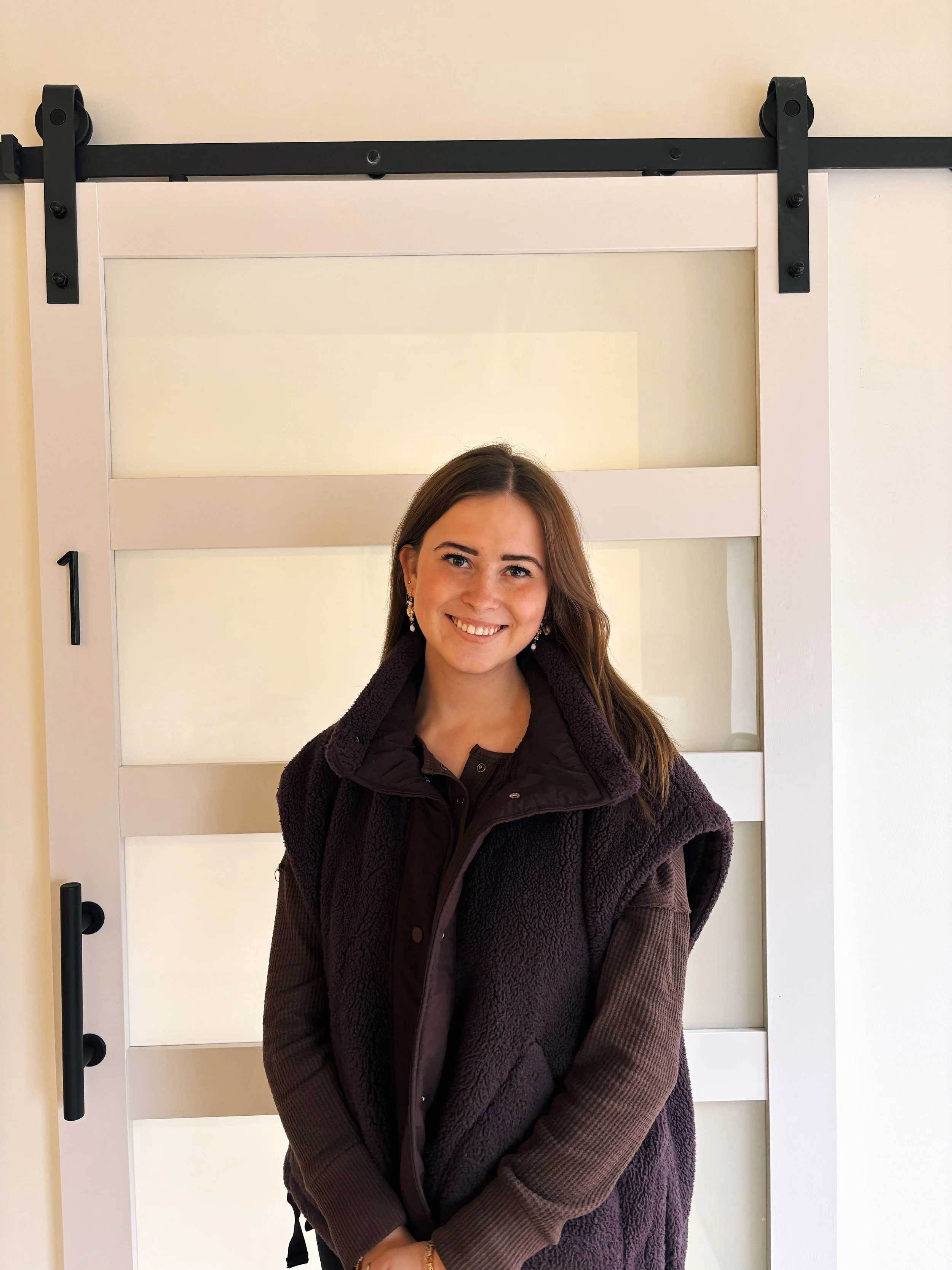 A woman with long brown hair wearing a black jacket and earrings smiling in front of a white sliding barn door.