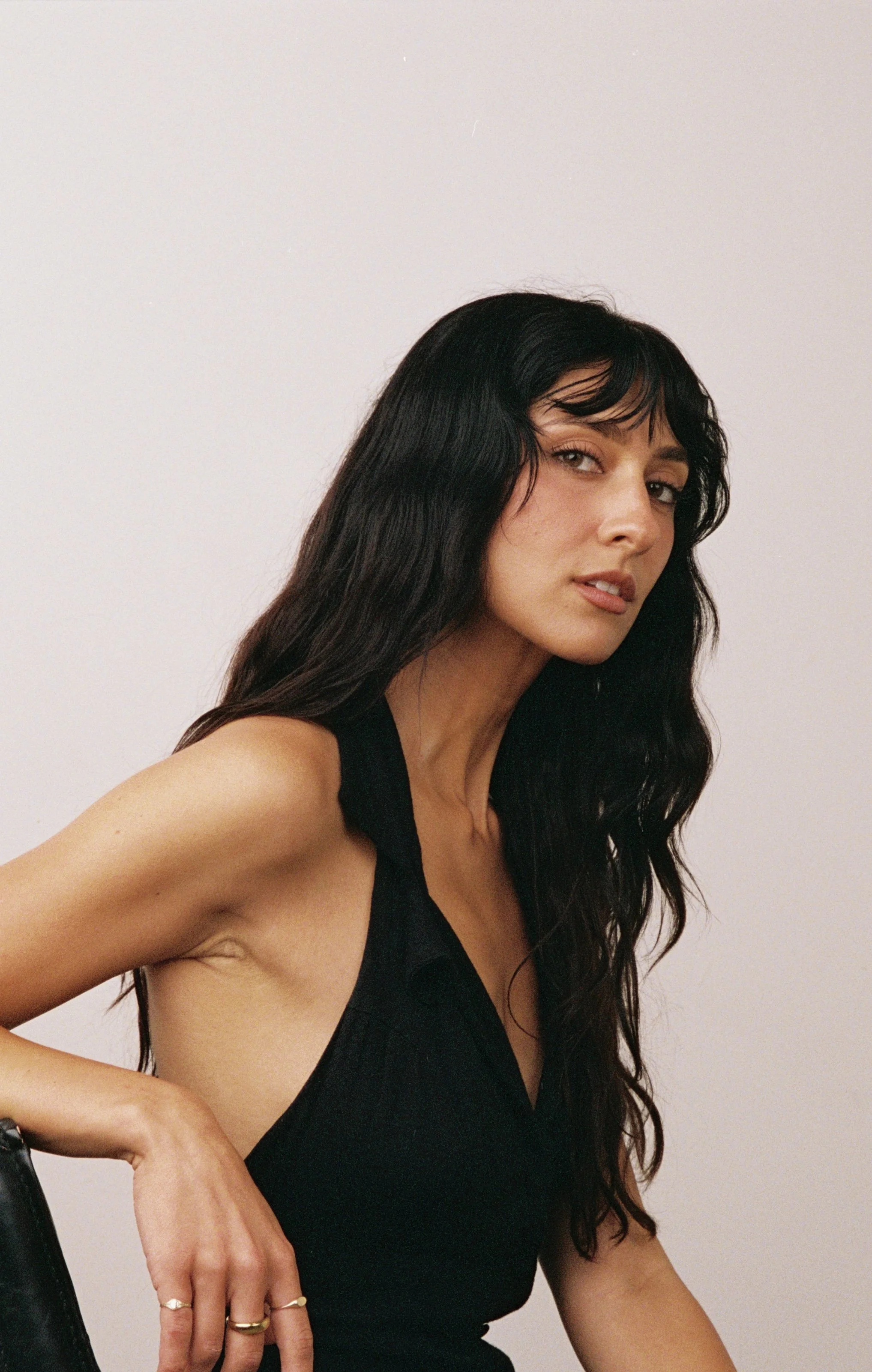 A woman with long, wavy black hair, wearing a black sleeveless top, posing against a plain white wall.