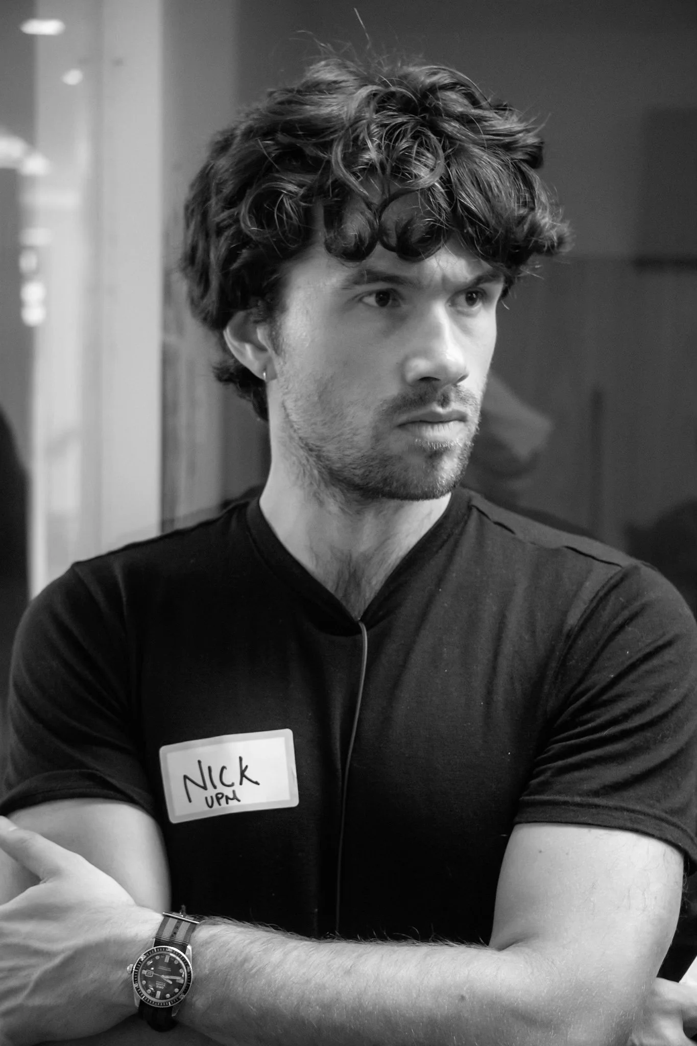 A young man with curly hair and a serious expression, wearing a dark shirt with a name tag that reads 'Nick' and 'UPM,' is sitting at a table with his arms crossed. He has a watch on his left wrist and appears to be at an indoor social or professional event.