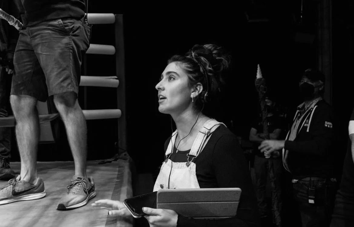 A woman with dark hair in a messy bun, wearing overalls and a black shirt, appears to be speaking or commanding on set. She holds a tablet and a phone. A person with shorts and sneakers stands on a stage or platform, with others in the background, some wearing masks.