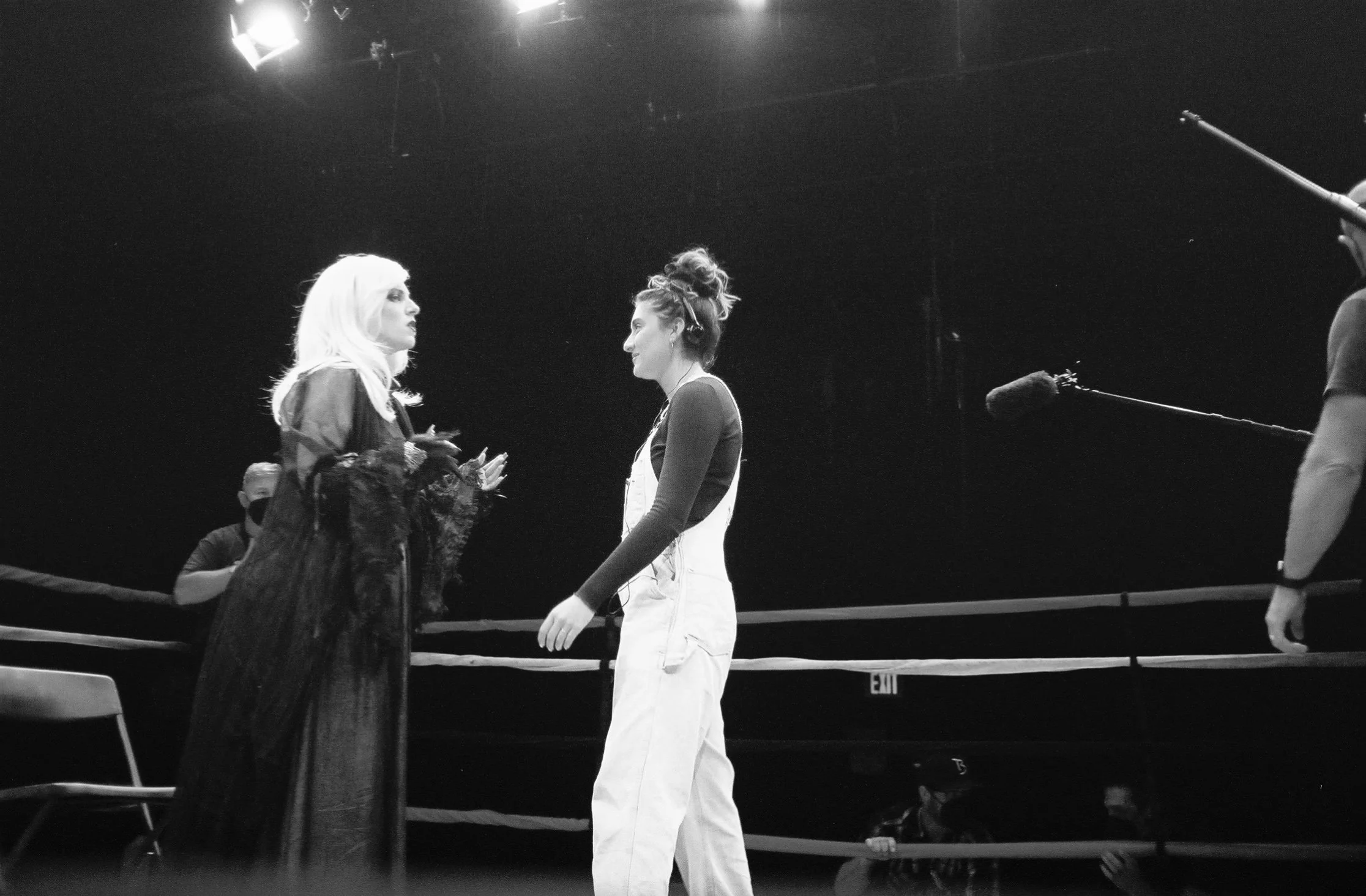 Two women are standing on a stage, engaged in a conversation, with stage lights above them. One woman has long, light-colored hair, and the other has dark, curly hair tied up. There are microphones and a chair visible on the stage.