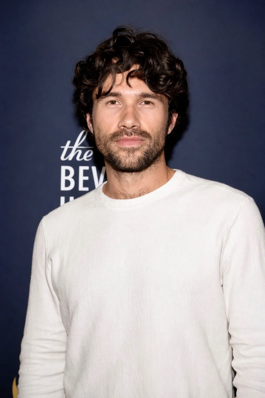 A man with dark curly hair, beard, and mustache wearing a white long-sleeve shirt standing in front of a blue backdrop with white text.