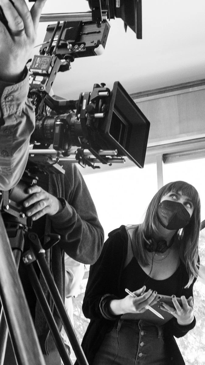 A woman wearing a face mask and headphones, holding a smartphone and pen, standing next to a camera operator using professional filming equipment.