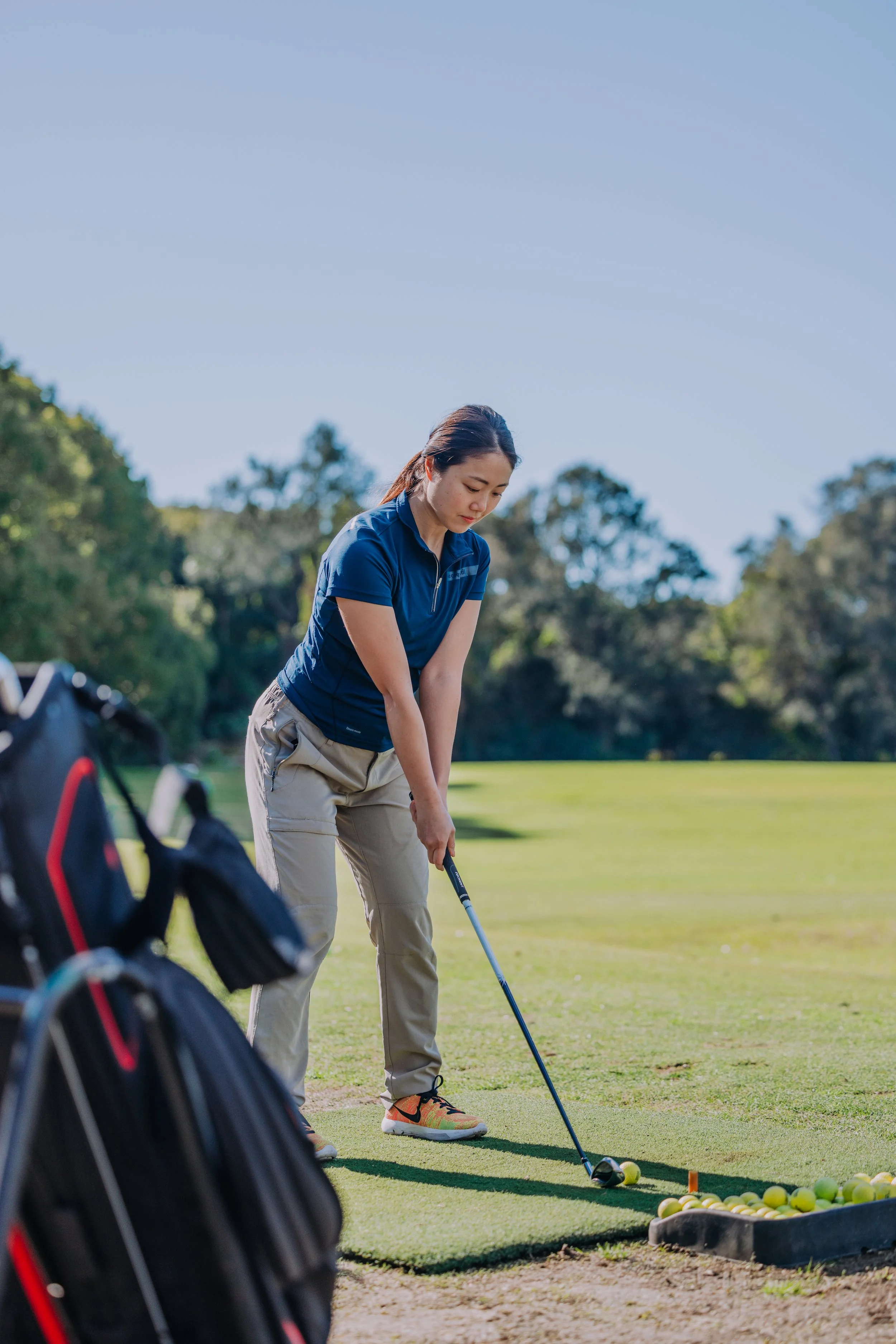 A woman in a blue shirt and khaki pants practicing her golf swing on a sunny day at a golf course, with a golf bag and golf balls nearby.