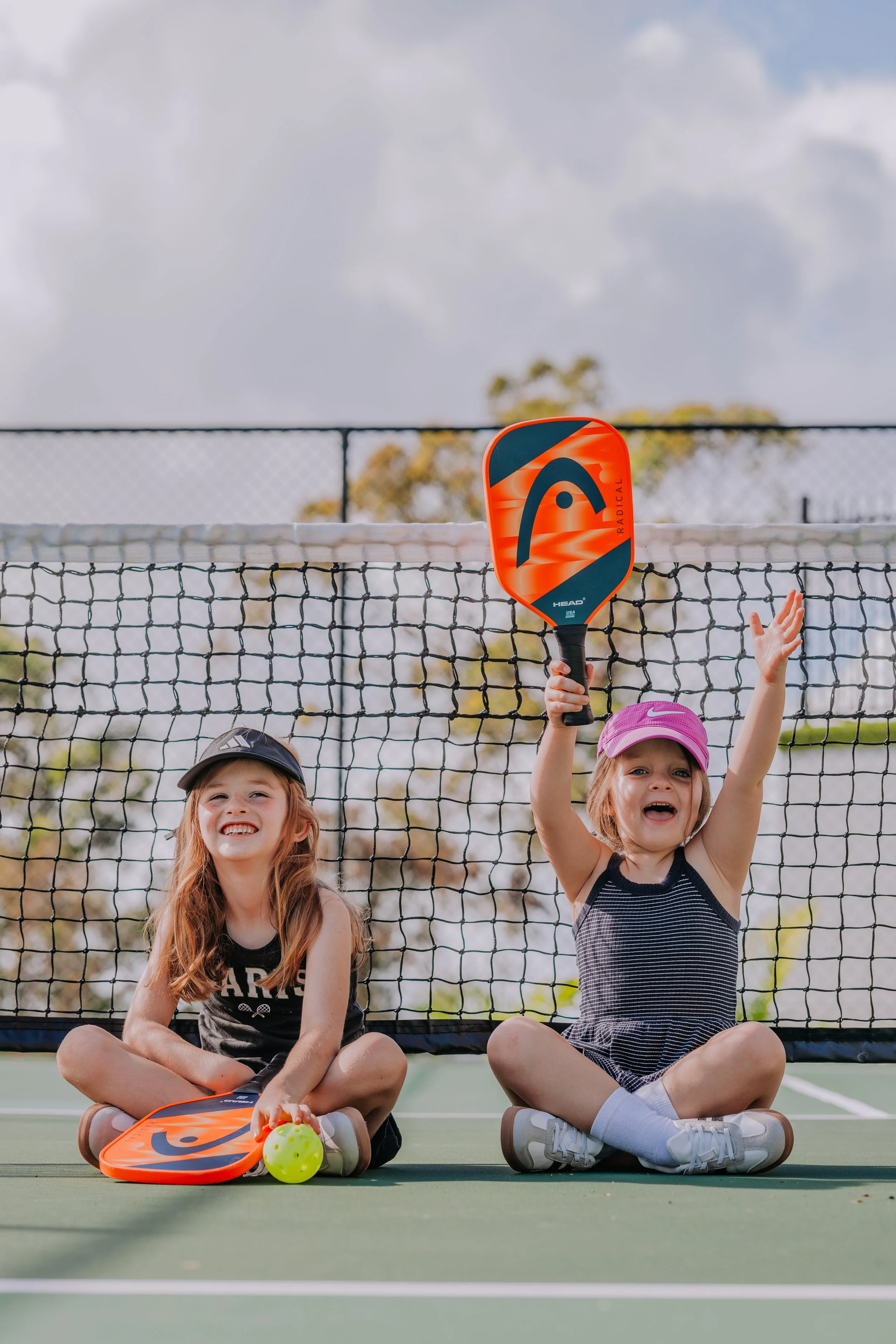 Two young girls sitting cross-legged on a tennis court behind the net, holding tennis paddles. The girl on the right is raising her paddle and smiling. Both girls are wearing casual sports clothes and caps, and one has a tennis ball and a paddle resting on her lap. The background shows a cloudy sky and trees.