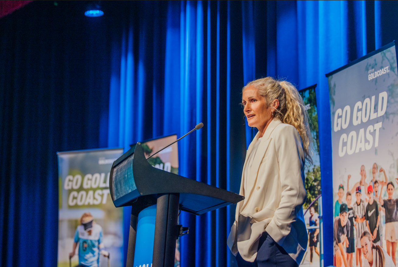 A woman in a white blazer speaking at a podium during a conference with blue curtains and colorful banners in the background that read "Go Gold Coast."